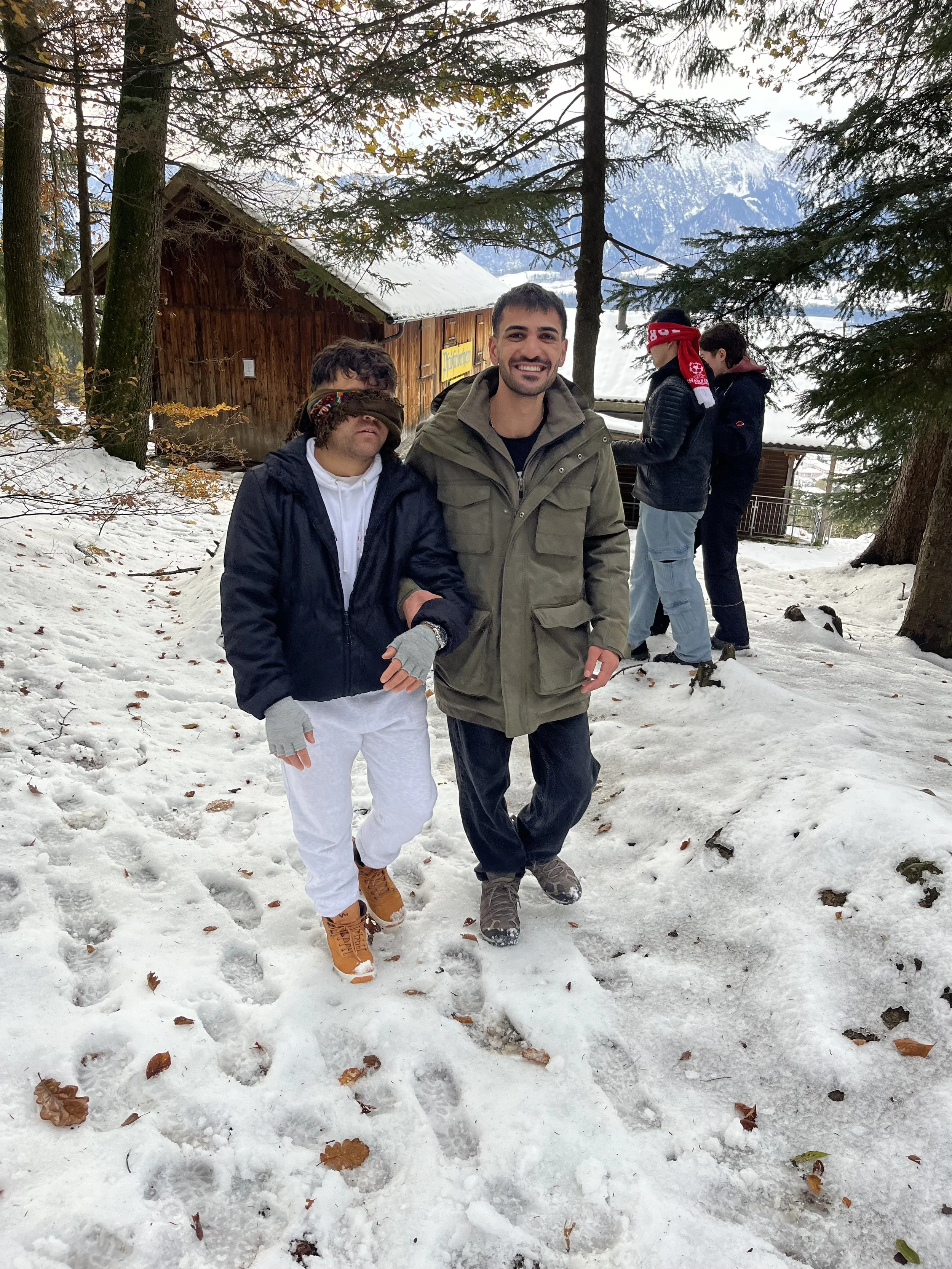 Drei Männer stehen im Schnee in einem Wald. Der Mann in der Mitte lächelt, die anderen beiden haben die Augen verbunden. Im Hintergrund sind Tannenbäume, eine Hütte und Berge sichtbar.