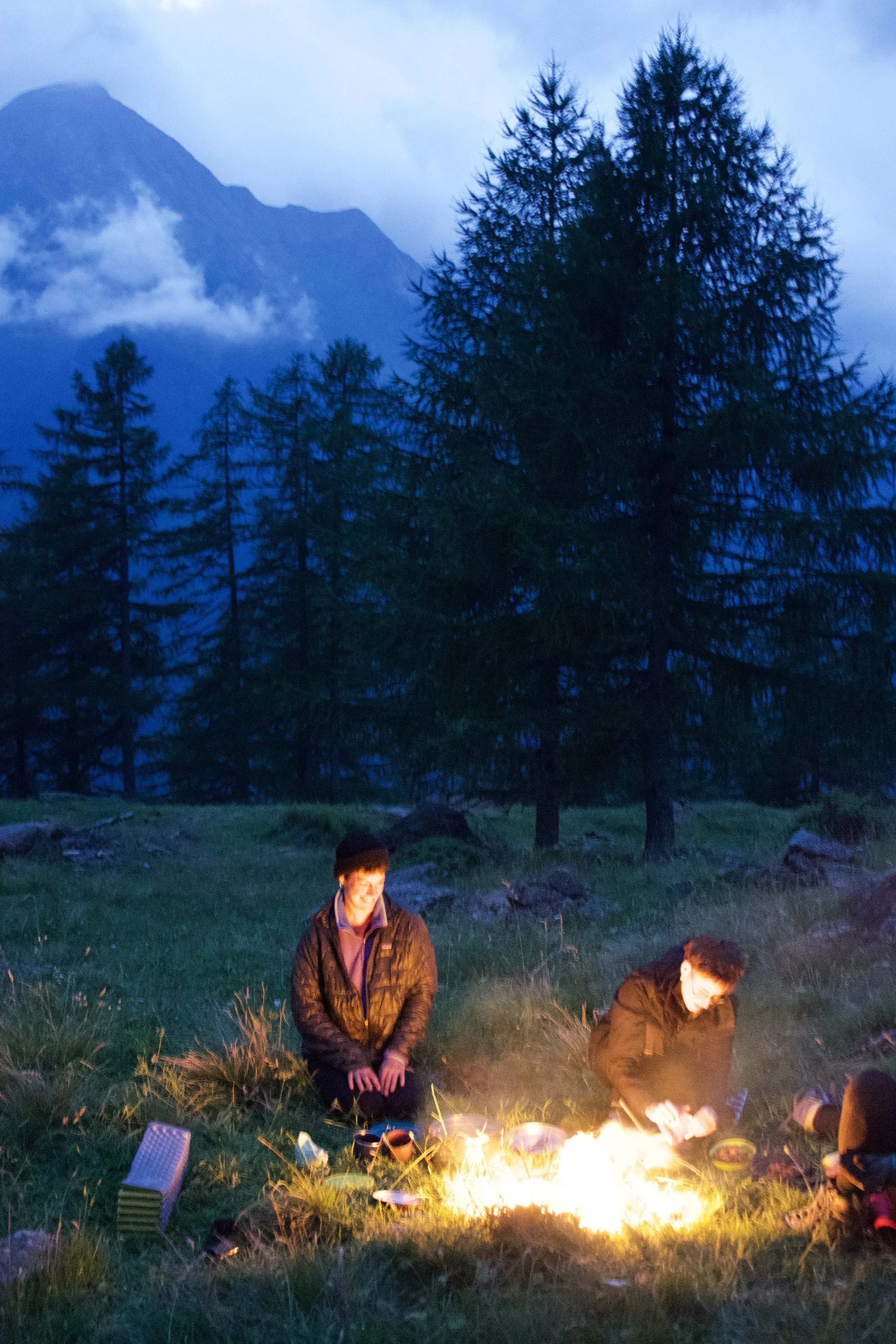 Zwei Personen sitzen an einem Lagerfeuer im Grünen, umgeben von Bäumen und Bergen bei Dämmerung.