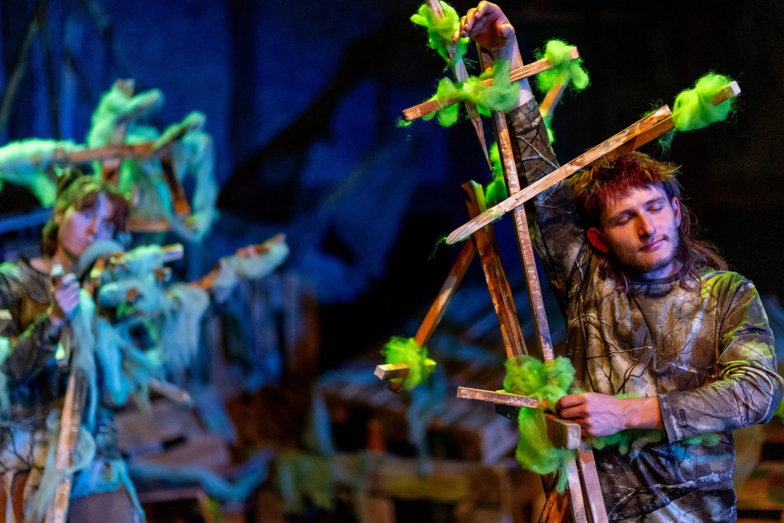 Zwei Schauspieler in der Theateraufführung, einer sitzt auf einer Leiter, der andere steht neben ihm. Beide tragen Kostüme mit grünem Filz und Holzstangen, in einer Szenerie, die eine Fantasiewelt darstellt.