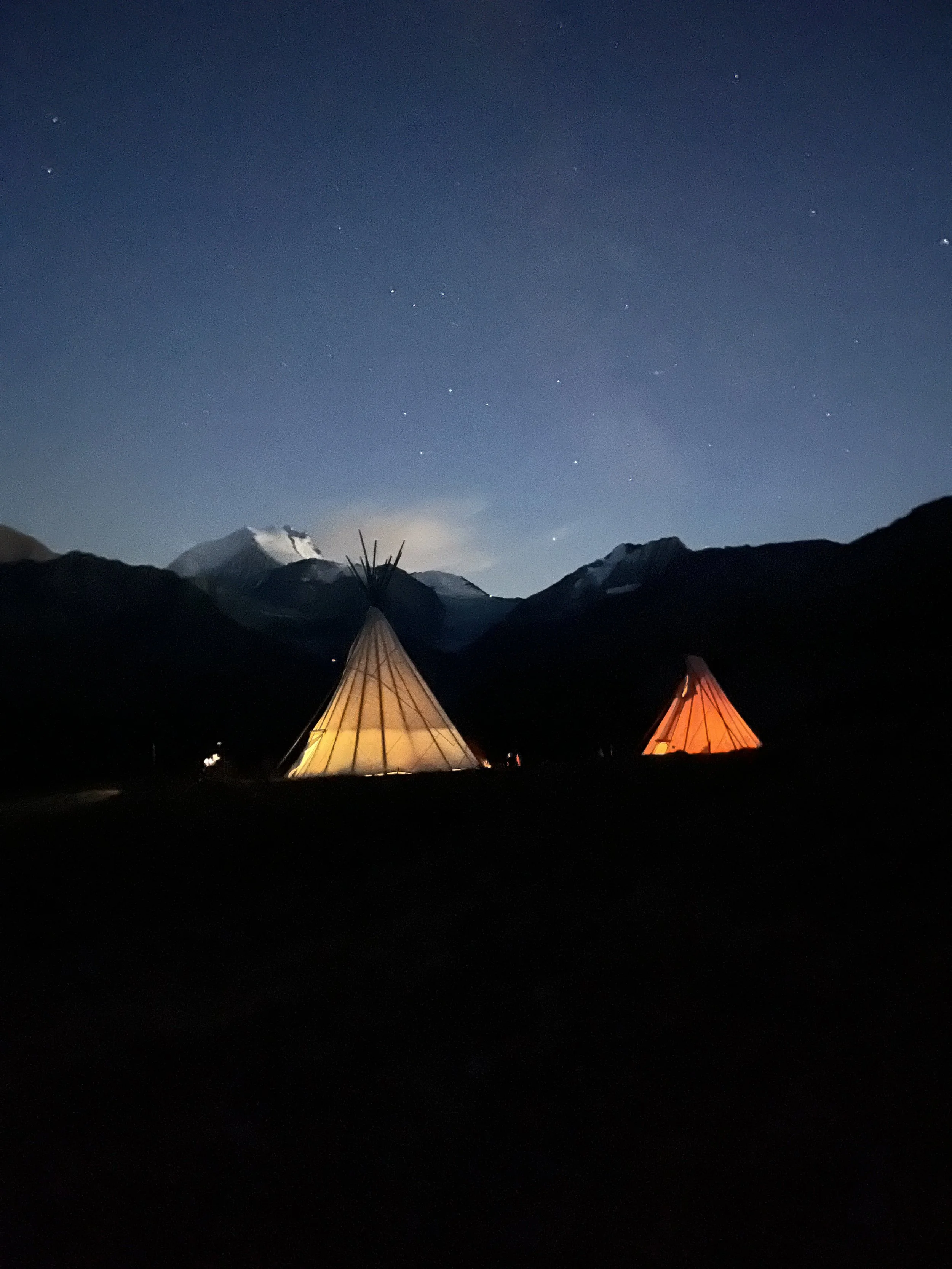 Zwei beleuchtete Tipis vor einer Bergkulisse bei Nacht, der Himmel ist sternenklar.