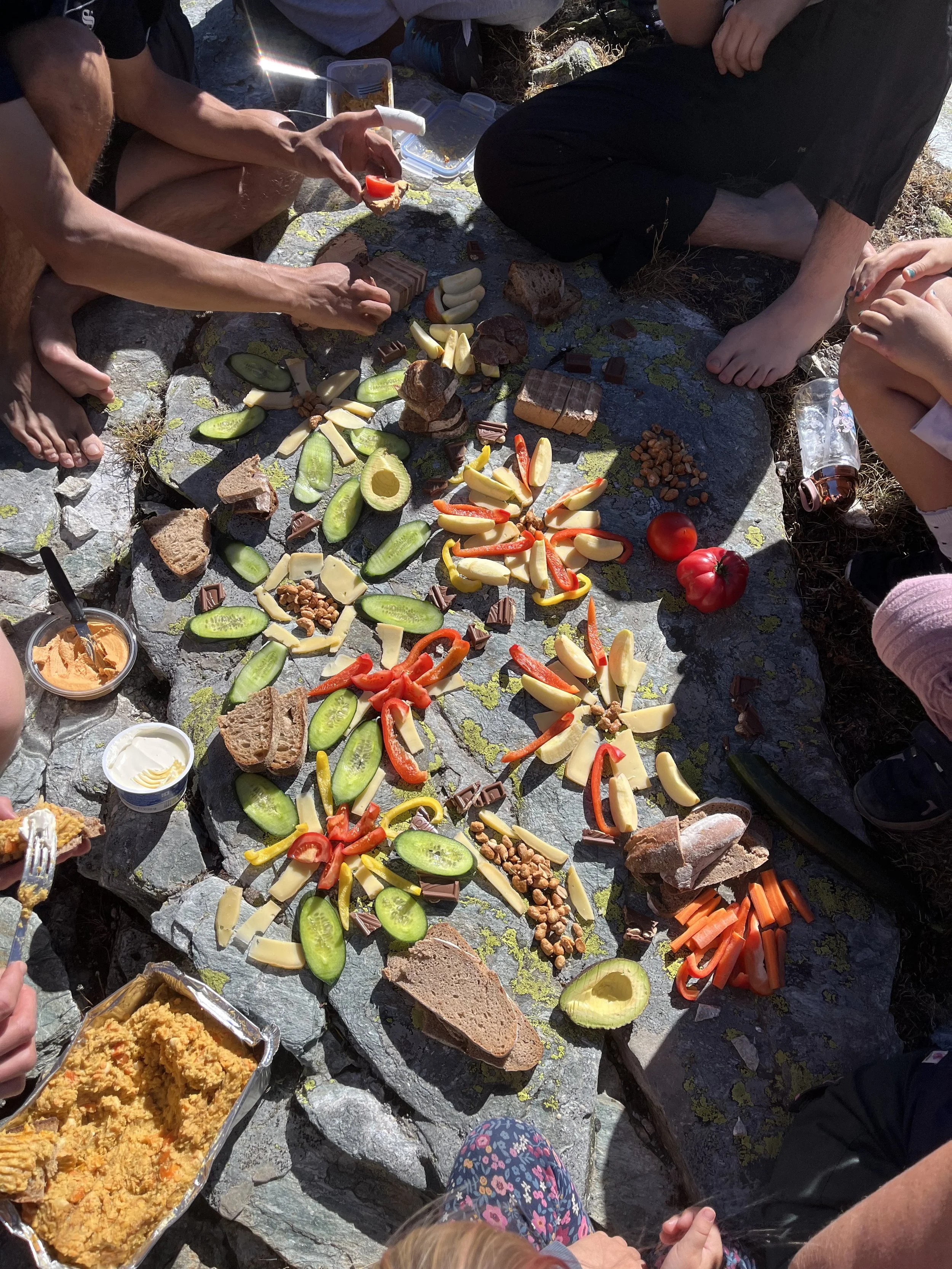 Gruppierung von Lebensmitteln, darunter Gurkenscheiben, Paprika, Käse, Nüsse, Brot und Tomaten, die auf einem Felsen für ein Picknick im Freien angeordnet sind.