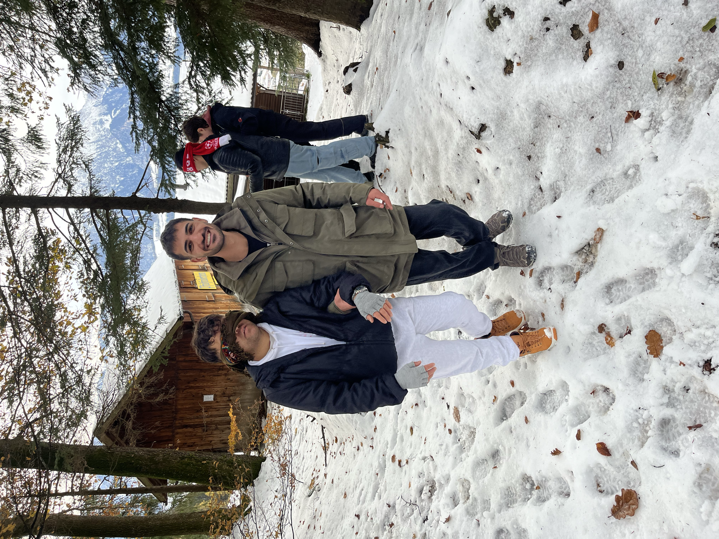 Fünf Menschen stehen im verschneiten Wald, zwei lachen, während sie die Kamera anschauen. Im Hintergrund sind Bäume und ein kleines Gebäude sichtbar.
