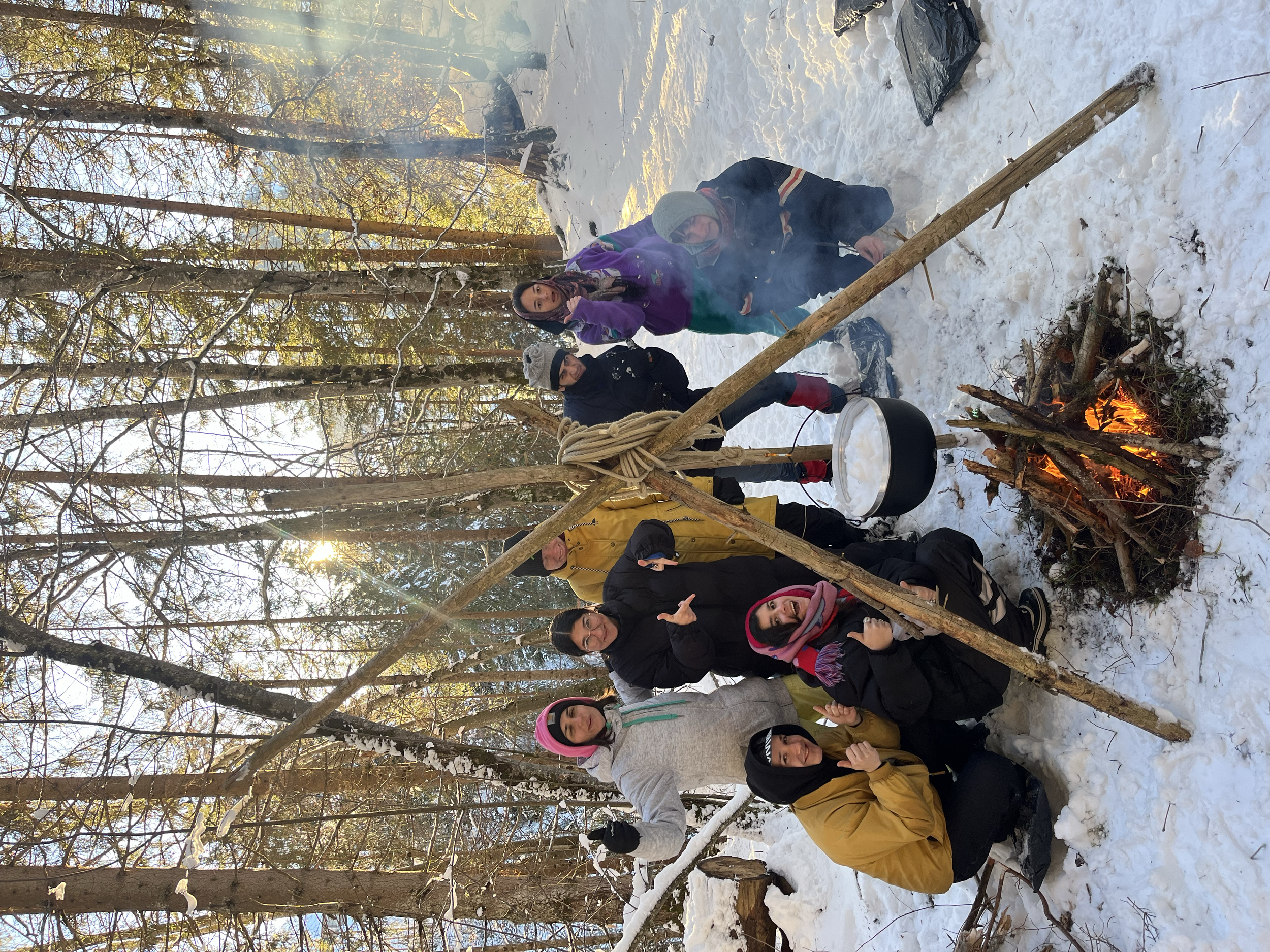 Eine Gruppe von Menschen in einem verschneiten Wald zündet ein Lagerfeuer an. Sie sind warm eingepackt und haben Campingausrüstung dabei.