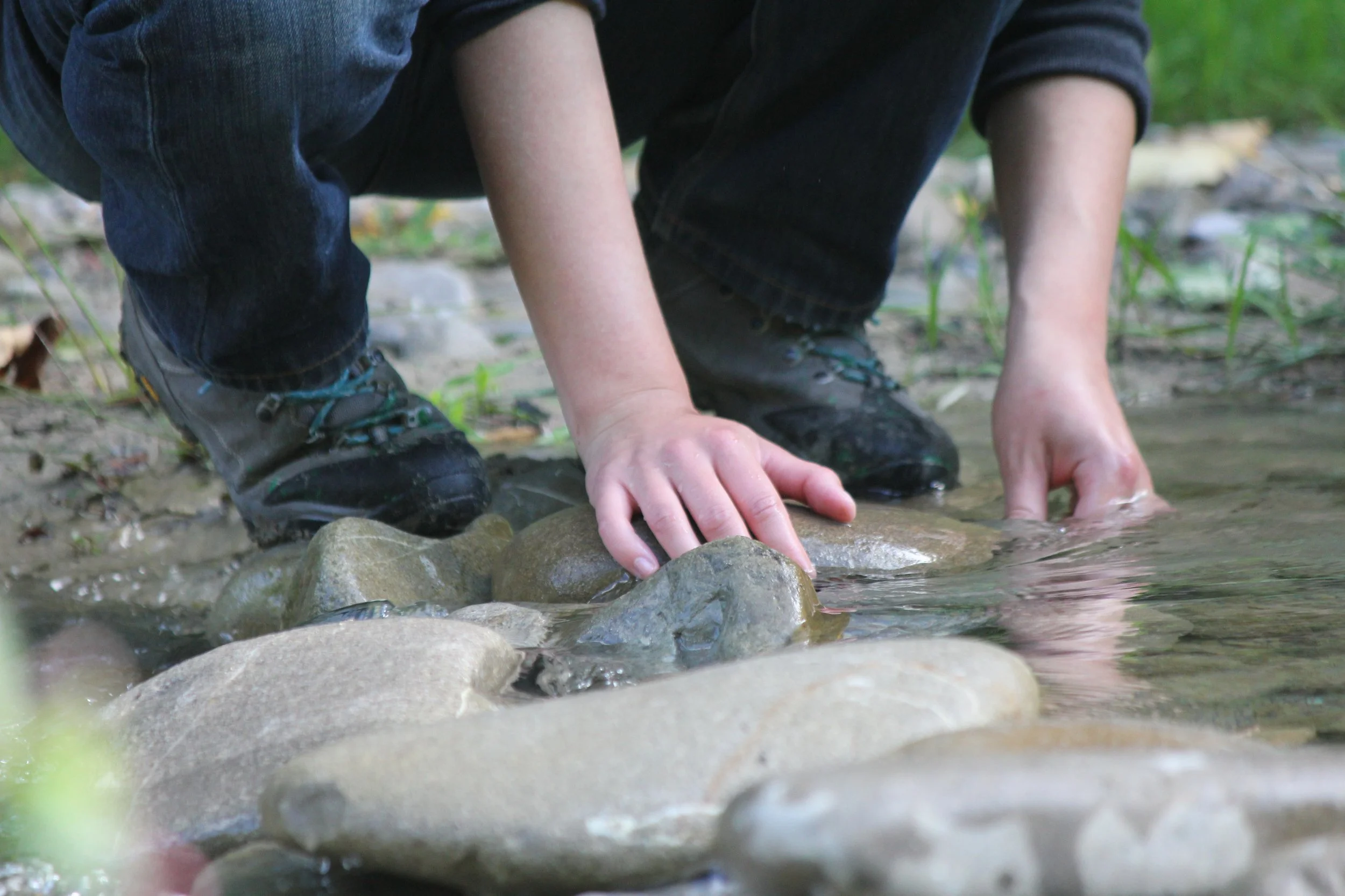 Kind kniet am Flussufer, berührt das Wasser mit den Händen, trägt Wanderschuhe, dunkle Kleidung, Naturumgebung mit Steinen und grünen Pflanzen.
