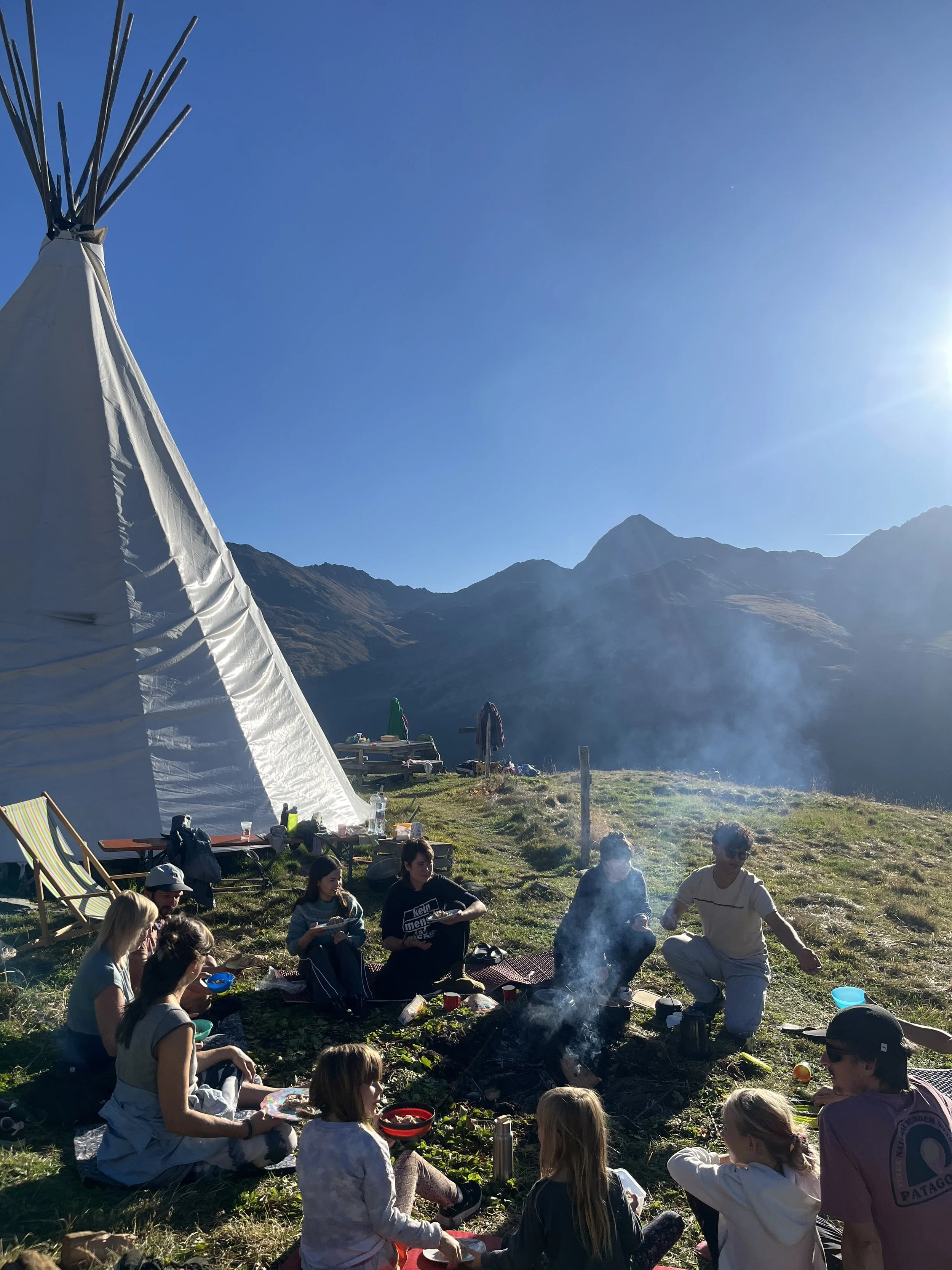 Gruppe von Menschen, darunter Kinder, die um ein Lagerfeuer in einer Berglandschaft sitzen, mit einem weißen Tipi-Zelt im Hintergrund bei sonnigem Wetter.