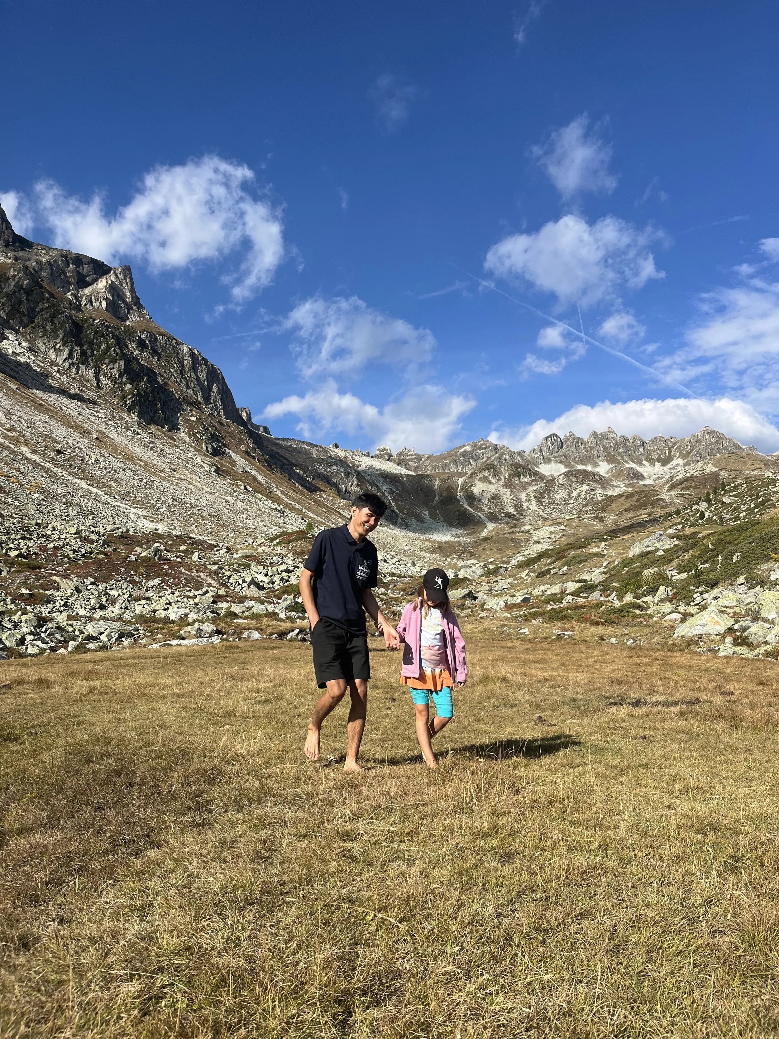 Ein Mann und ein Kind gehen barfuß durch ein offenes, grasbewachsenes Feld in den Bergen bei schönem Wetter, blauer Himmel mit wenigen Wolken.