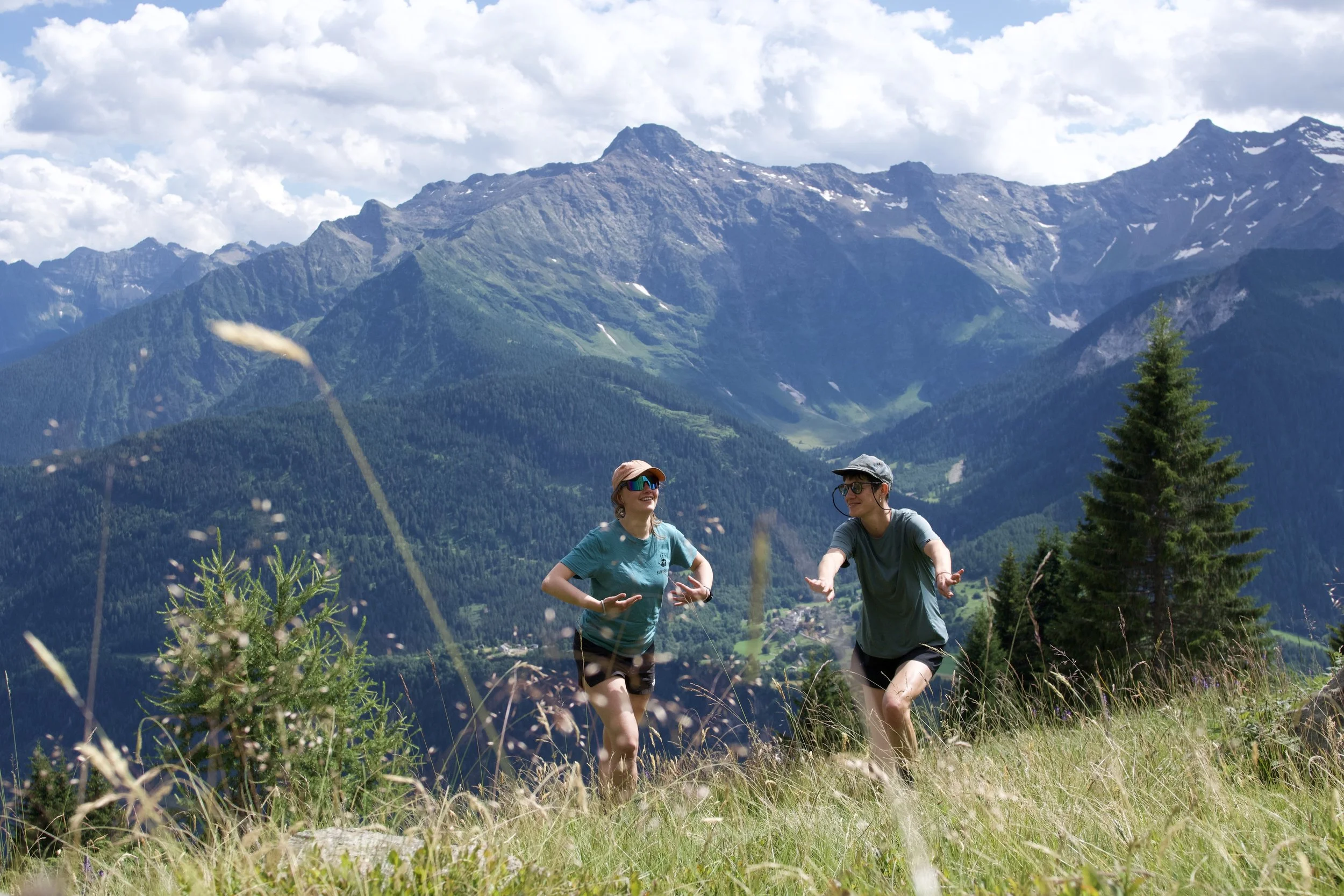 Zwei Menschen joggen in den Bergen, umgeben von grünen Wäldern und hohen Gipfeln, bei sonnigem Wetter.
