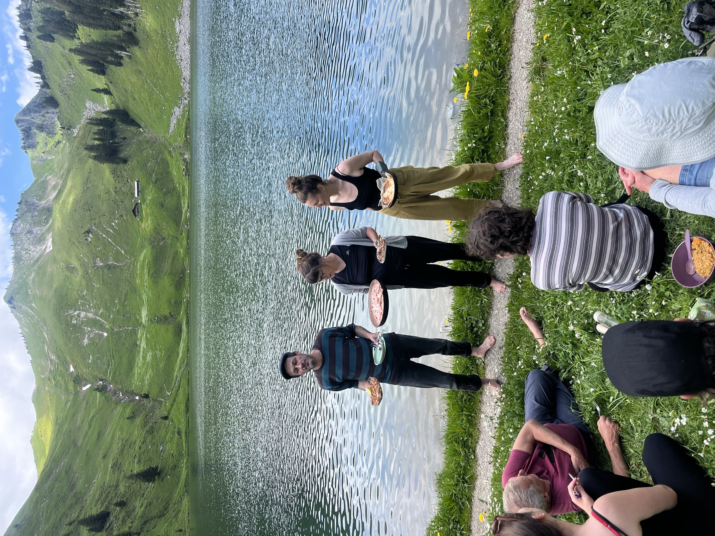 Menschen beim Picknick am Ufer eines Sees mit grünen Bergen im Hintergrund, einige sitzen auf dem Gras und andere stehen und tragen Teller mit Essen.