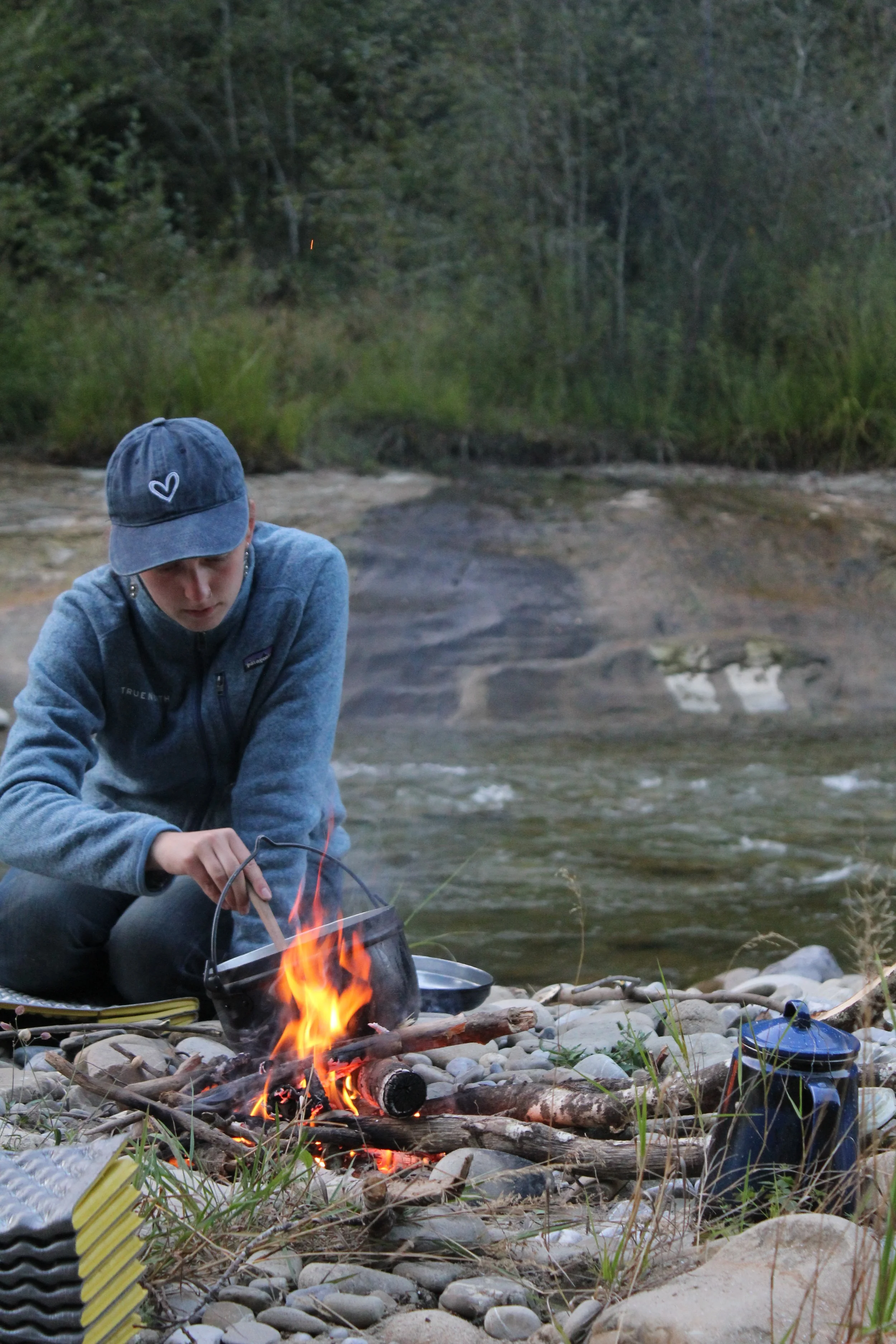 Junge Person, die an einem Lagerfeuer am Fluss in der Natur sitzt und kocht.