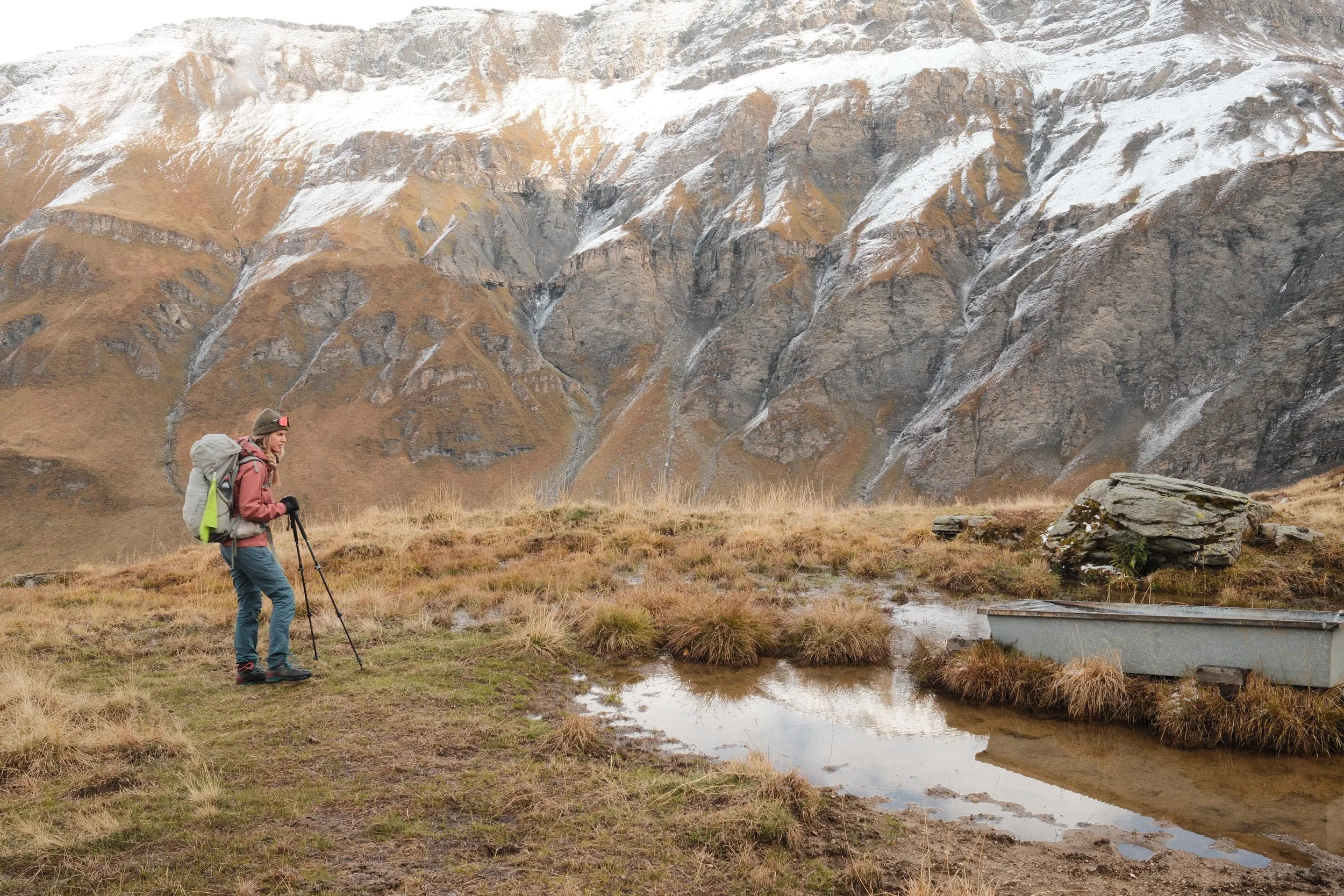 Eine Person beim Wandern in einer bergigen, kargen Landschaft mit Schnee auf den Gipfeln, trägt einen Rucksack und verwendet Wanderstöcke.