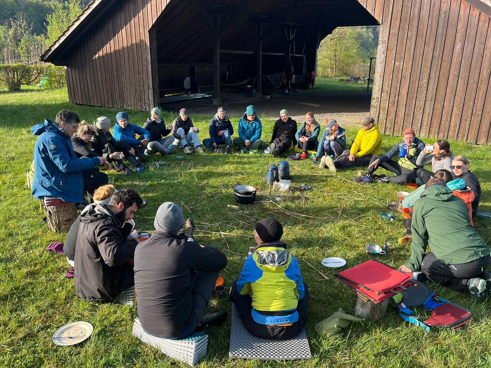 Eine Gruppe von Menschen sitzt im Kreis auf dem Gras vor einem Holzschuppen, beim Camping. Sie sind in wetterfester Kleidung, einige tragen Hüte und Sonnenbrillen. Es gibt Tassen, Teller und Campingausrüstung auf dem Boden, die Sonne scheint, und es ist grün und ländlich im Hintergrund.