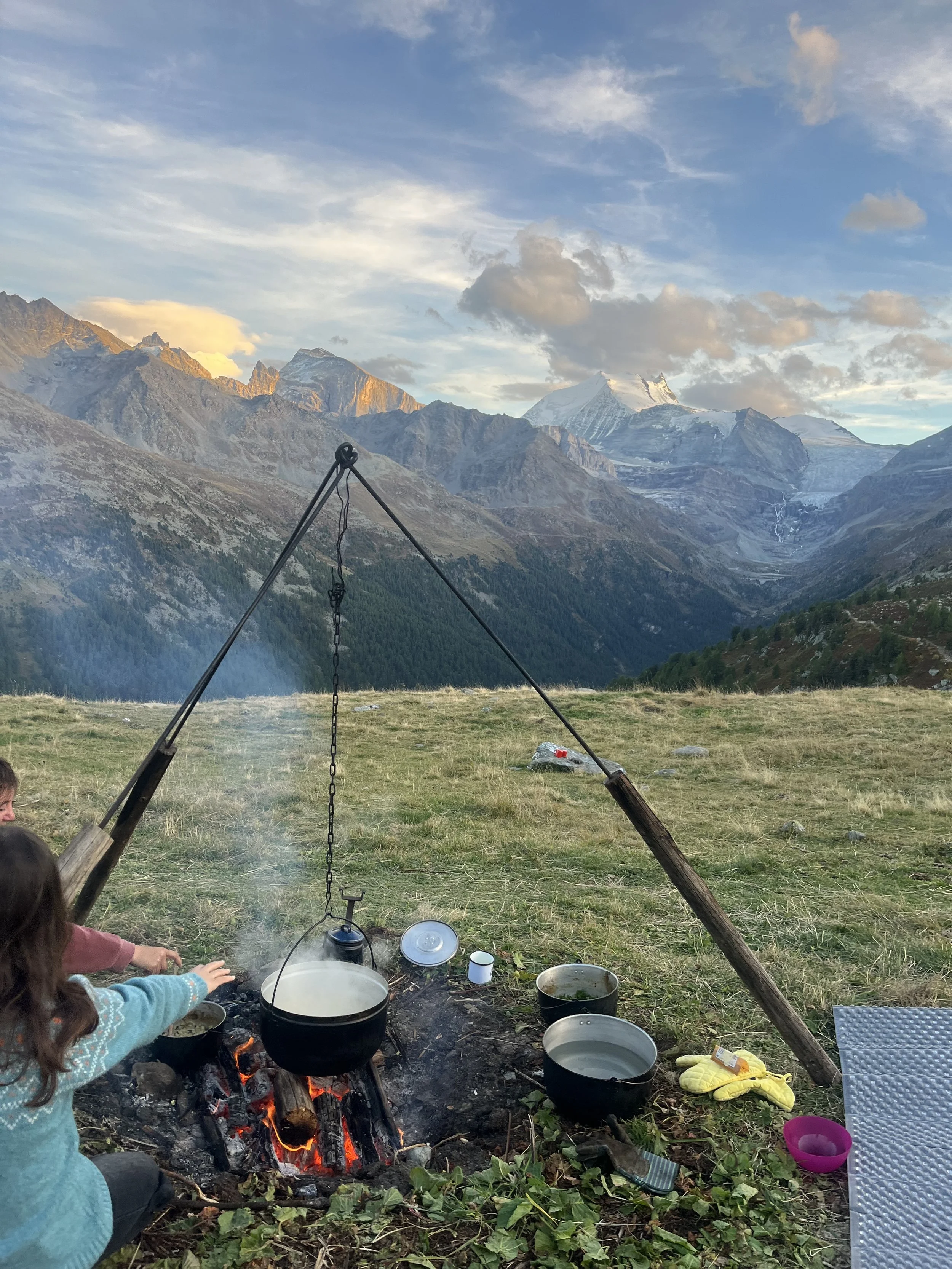 Zwei Kinder beim Kochen über einem Feuer in einer bergigen, grünen Landschaft mit hohen Bergen im Hintergrund.