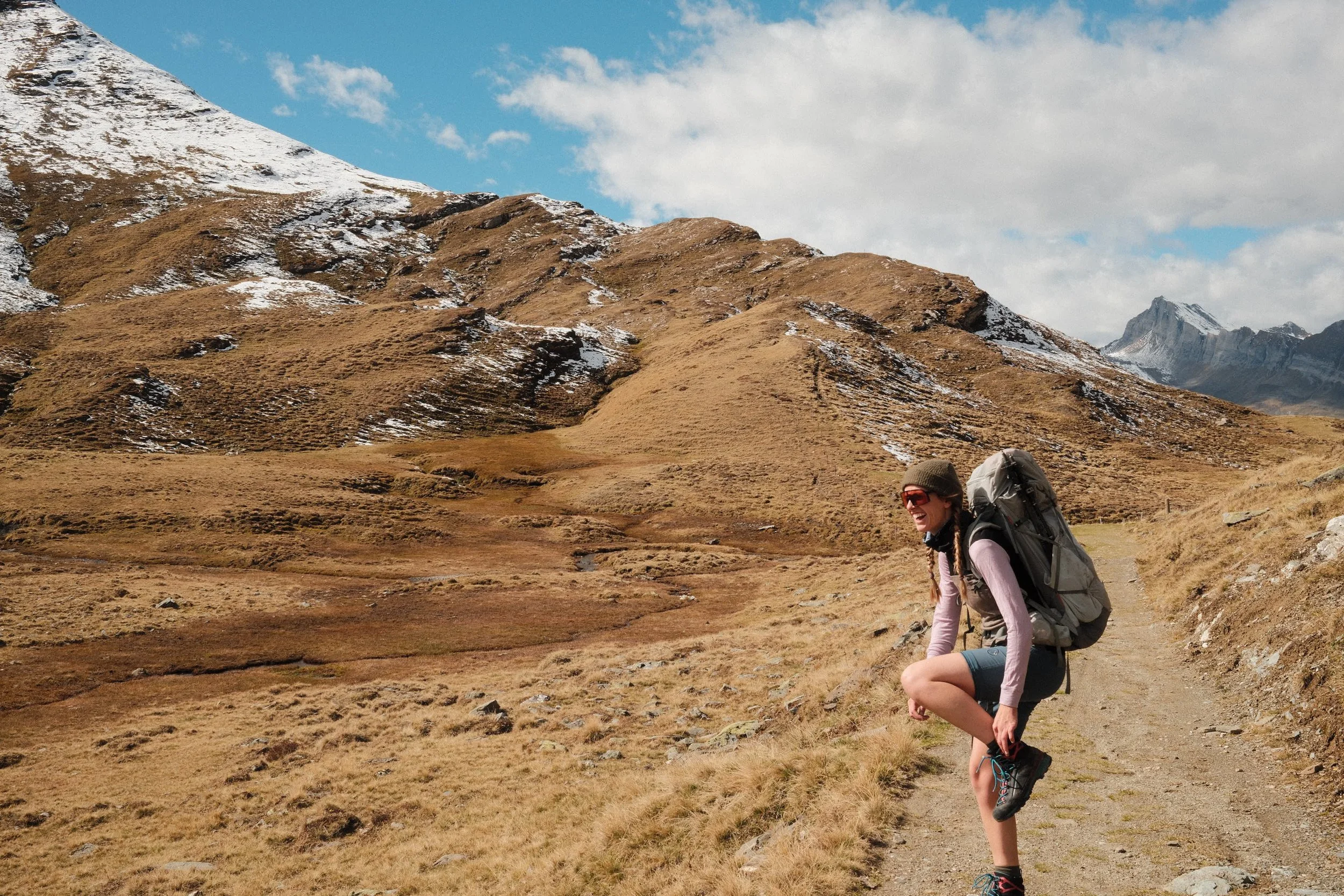 Junge Frau mit Rucksack beim Wandern in den Bergen, lachen, sonniger Tag, trockene Graslandschaft, Schnee auf den Bergen, blaue Wolken am Himmel.