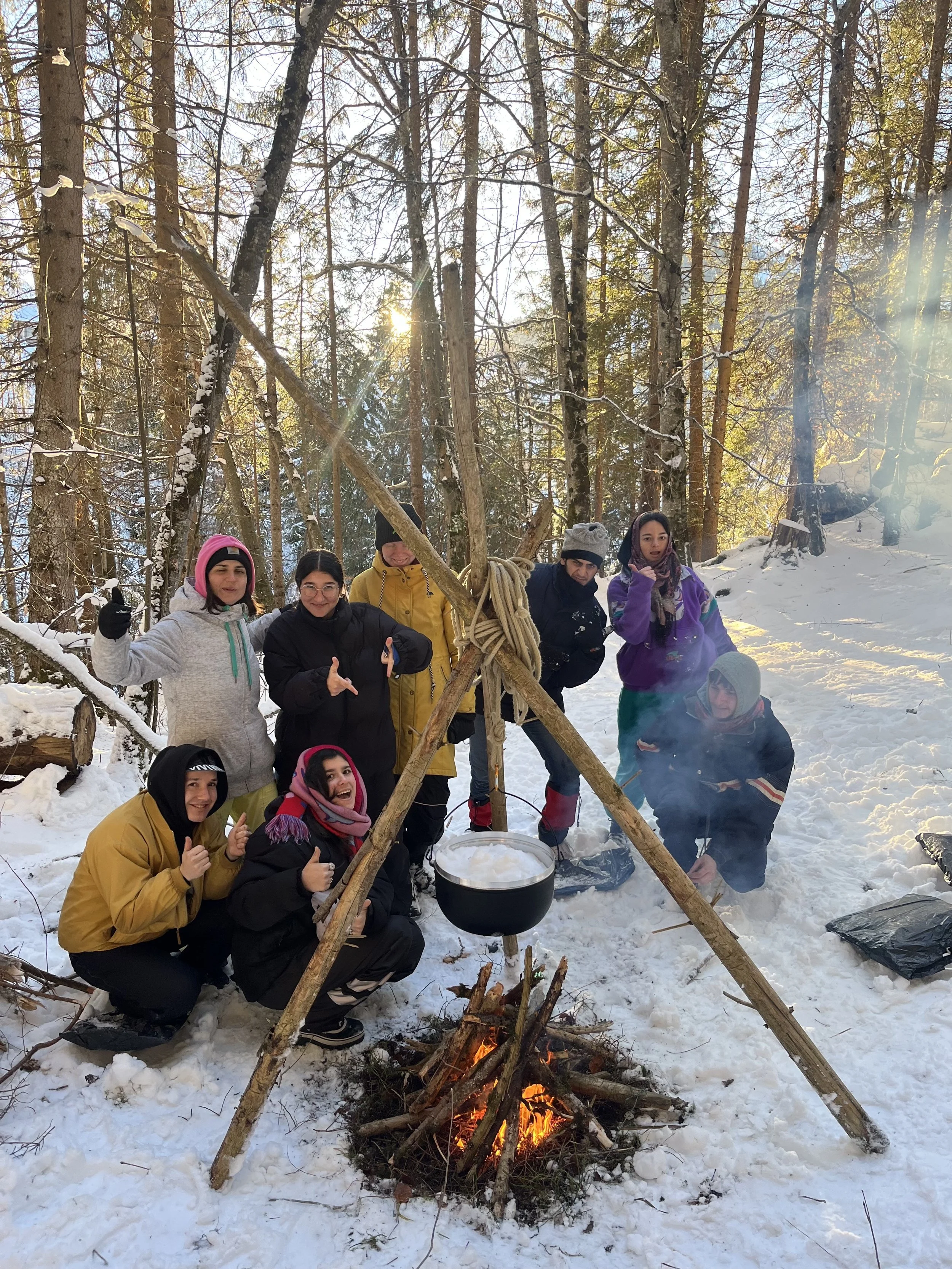 Gruppe von acht Personen beim Kochen über einem Lagerfeuer in einem verschneiten Wald, einige machen Daumen hoch, andere schauen in die Kamera, ein Topf hängt an einer improvisierten Unterkonstruktion aus Holzstücken.