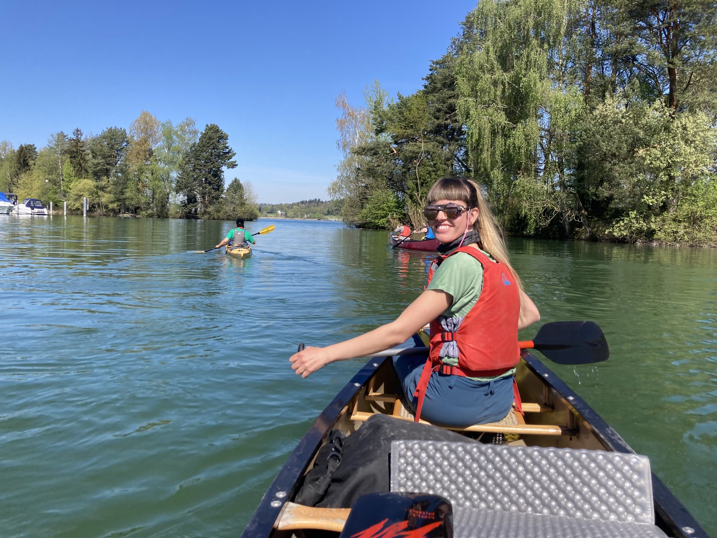 Junge Frau in einem Kajak auf einem Fluss, lächelnd, trägt Sonnenbrille und eine rote Schwimmweste, umgeben von grünen Bäumen bei sonnigem Wetter.