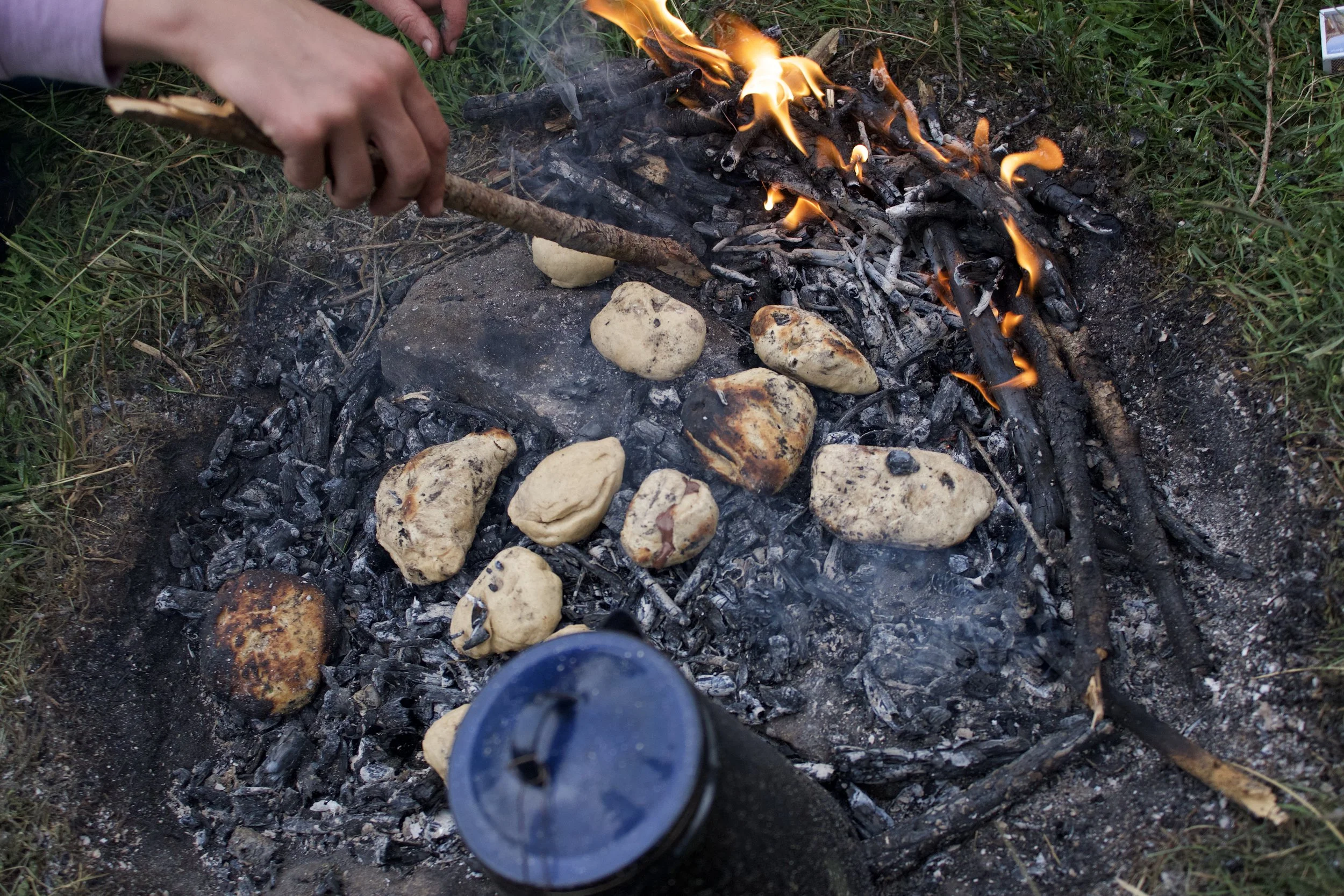 Person beim Kochen über offenem Feuer mit Steinen und Brötchen, umliegendes Gras.