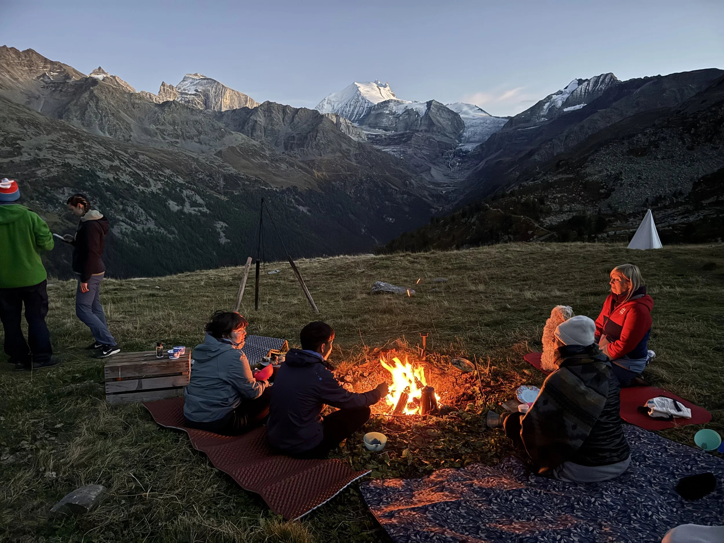 Menschen sitzen um ein Lagerfeuer auf einer Wiese in einer Bergregion bei Sonnenuntergang, im Hintergrund sind hohe, schneebedeckte Berge.