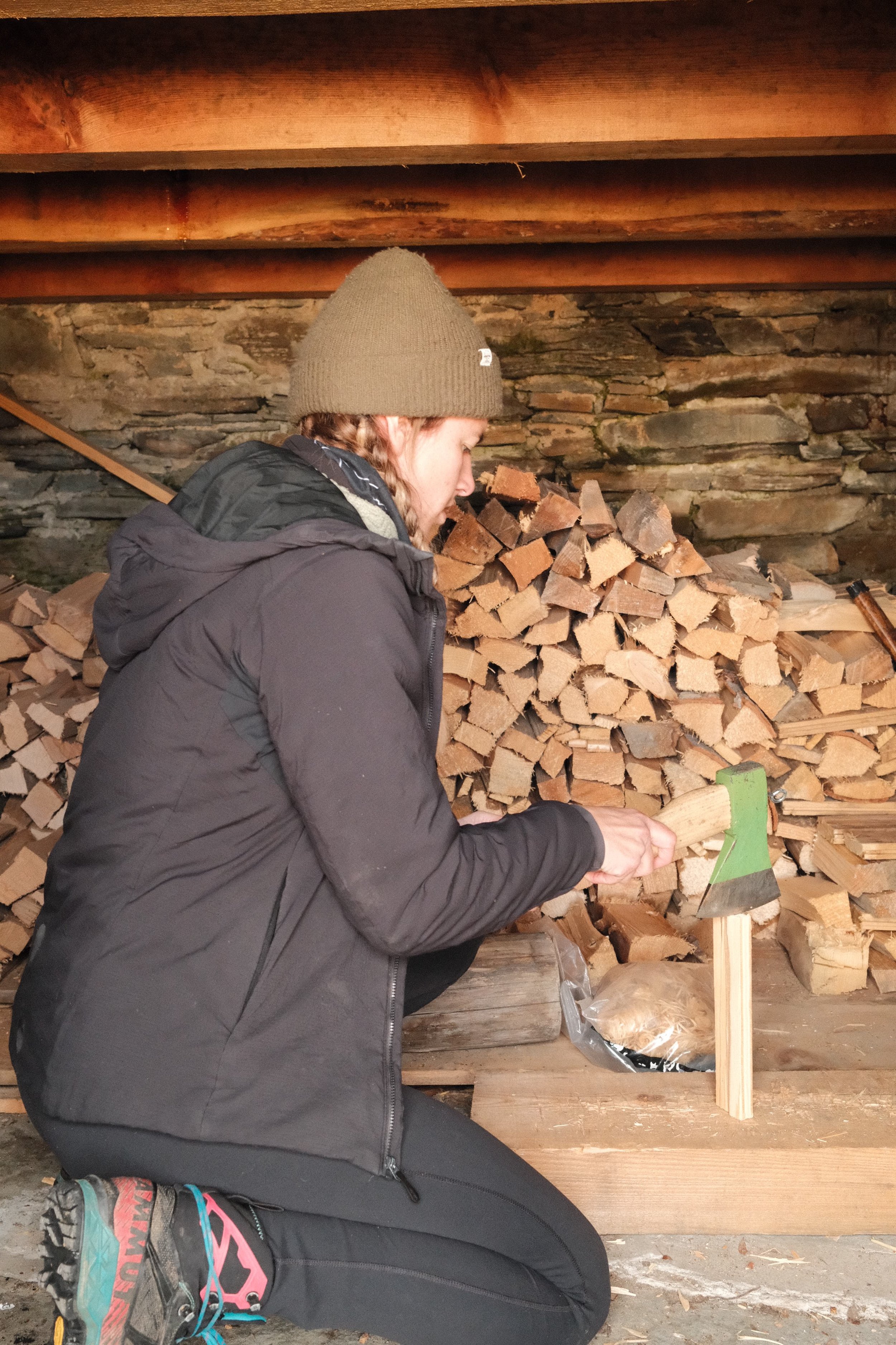 Eine Frau trägt eine schwarze Jacke und eine braune Mütze, sie schwingt eine Axt, um Holz zu zerkleinern, im Hintergrund viele Holzscheite.
