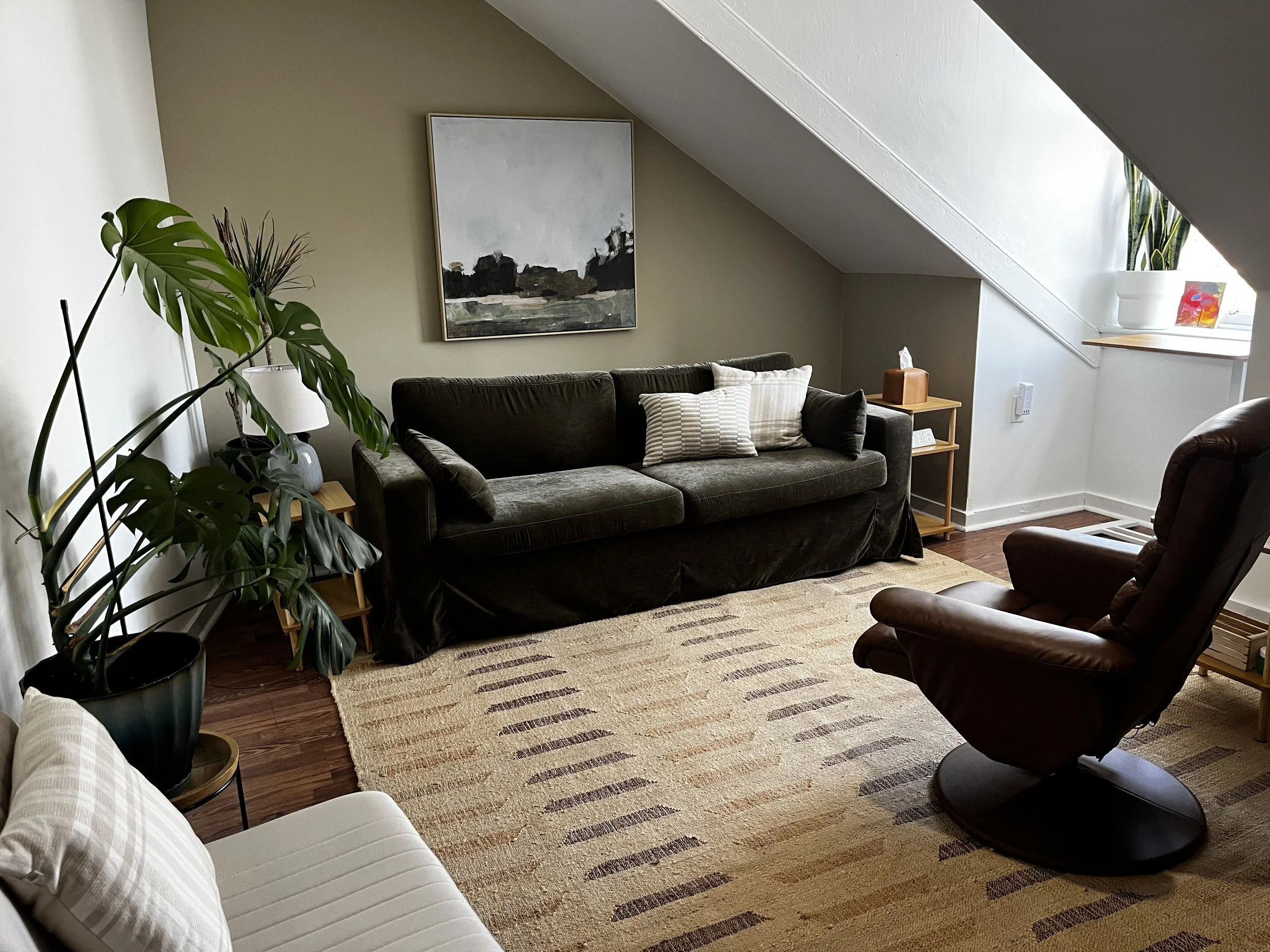 The Luna Therapy Group office space with a comfortable dark green sofa, a brown leather chair, a large potted plant, a beige area rug, a painting on the wall, and a window with a plant on the windowsill.