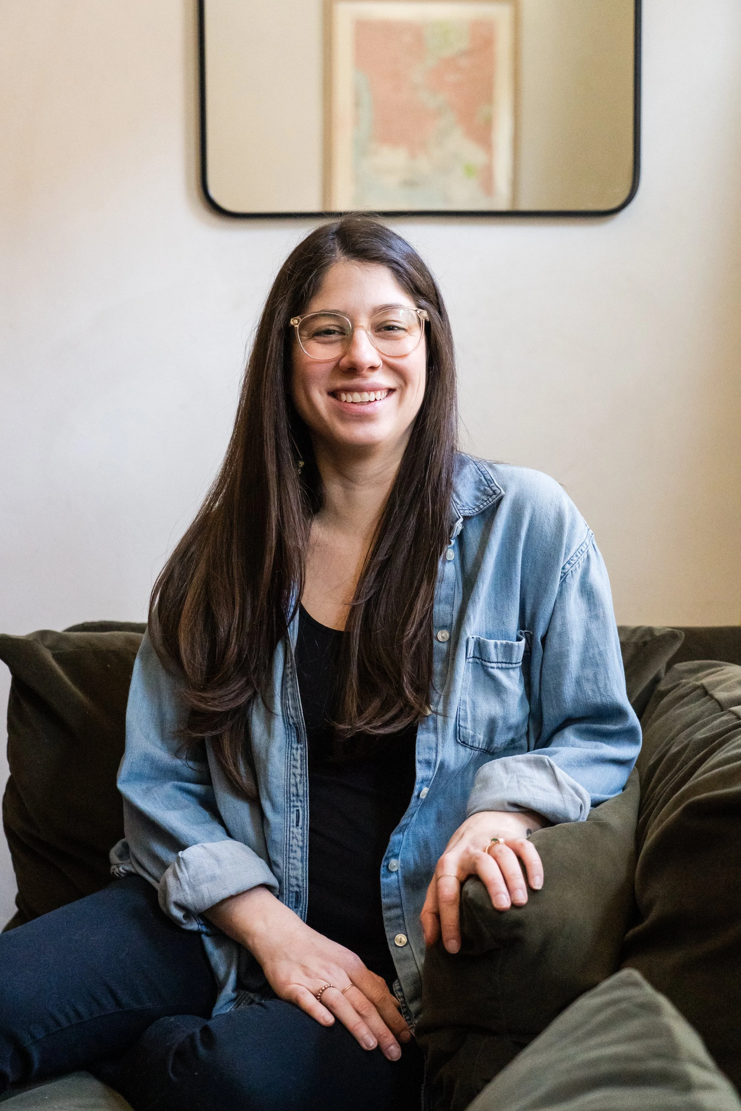 A kind female clinician, smiling with long dark hair and glasses, sits on a couch.