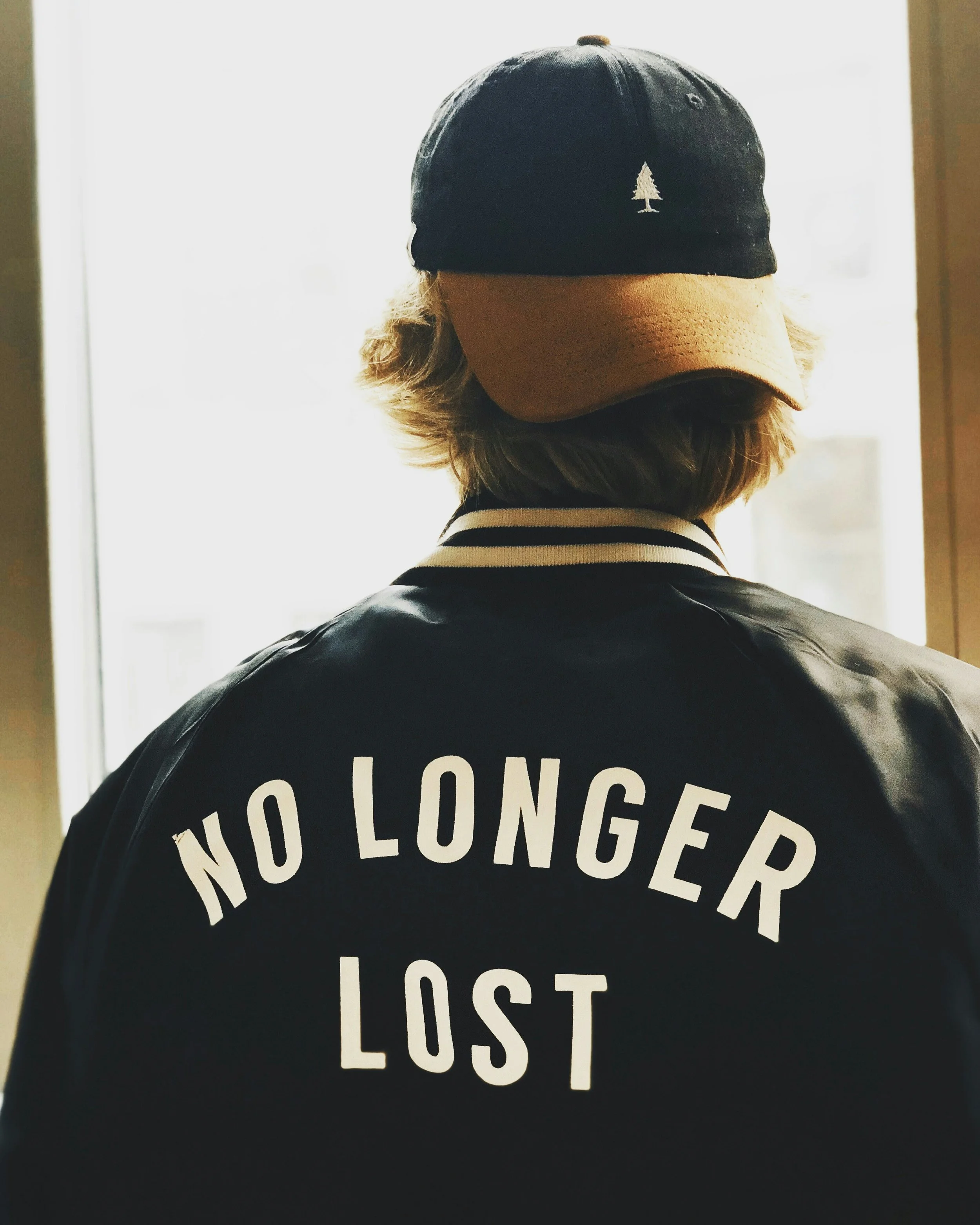 Back view of a person wearing a black and white varsity jacket with the words 'NO LONGER LOST' printed on the back, and a navy blue baseball cap with a tan brim and a white tree logo on the front.