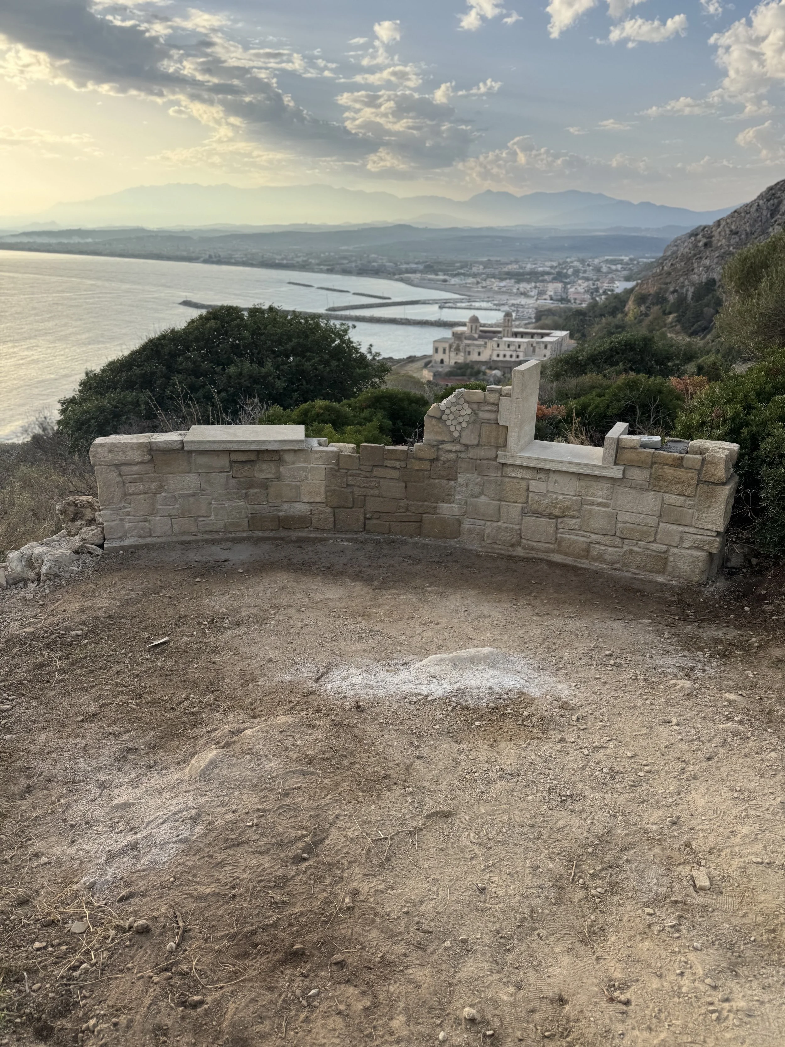 Blick von einem Aussichtspunkt auf das Meer, Berge und eine Stadt im Hintergrund, mit einer kleinen, halb fertig gebaut wirkenden Mauer im Vordergrund.