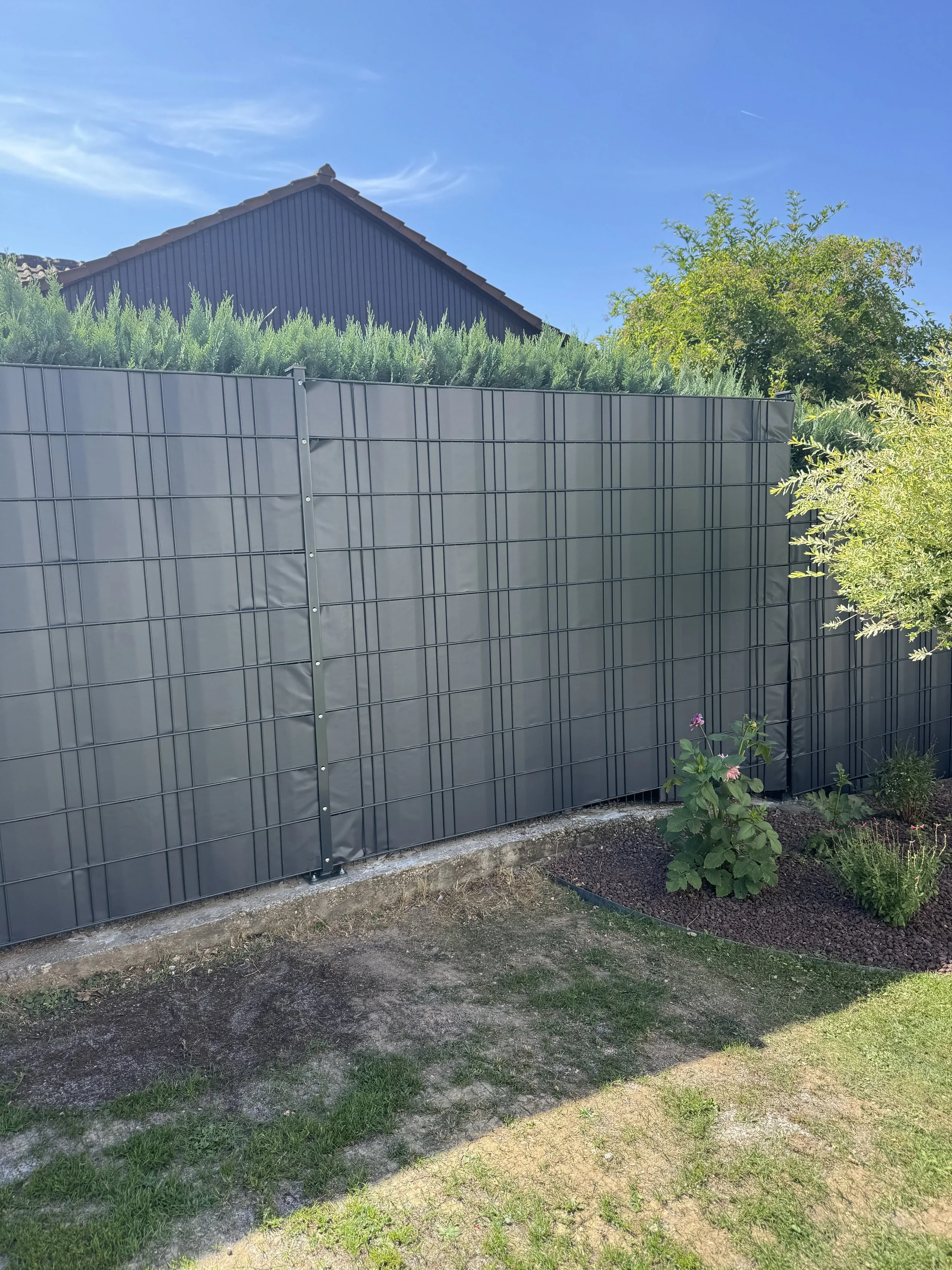 Garten mit grauem Metallzaun, blühende Pflanzen, Sand- und Grasflächen, blauem Himmel, Hausdach im Hintergrund.
