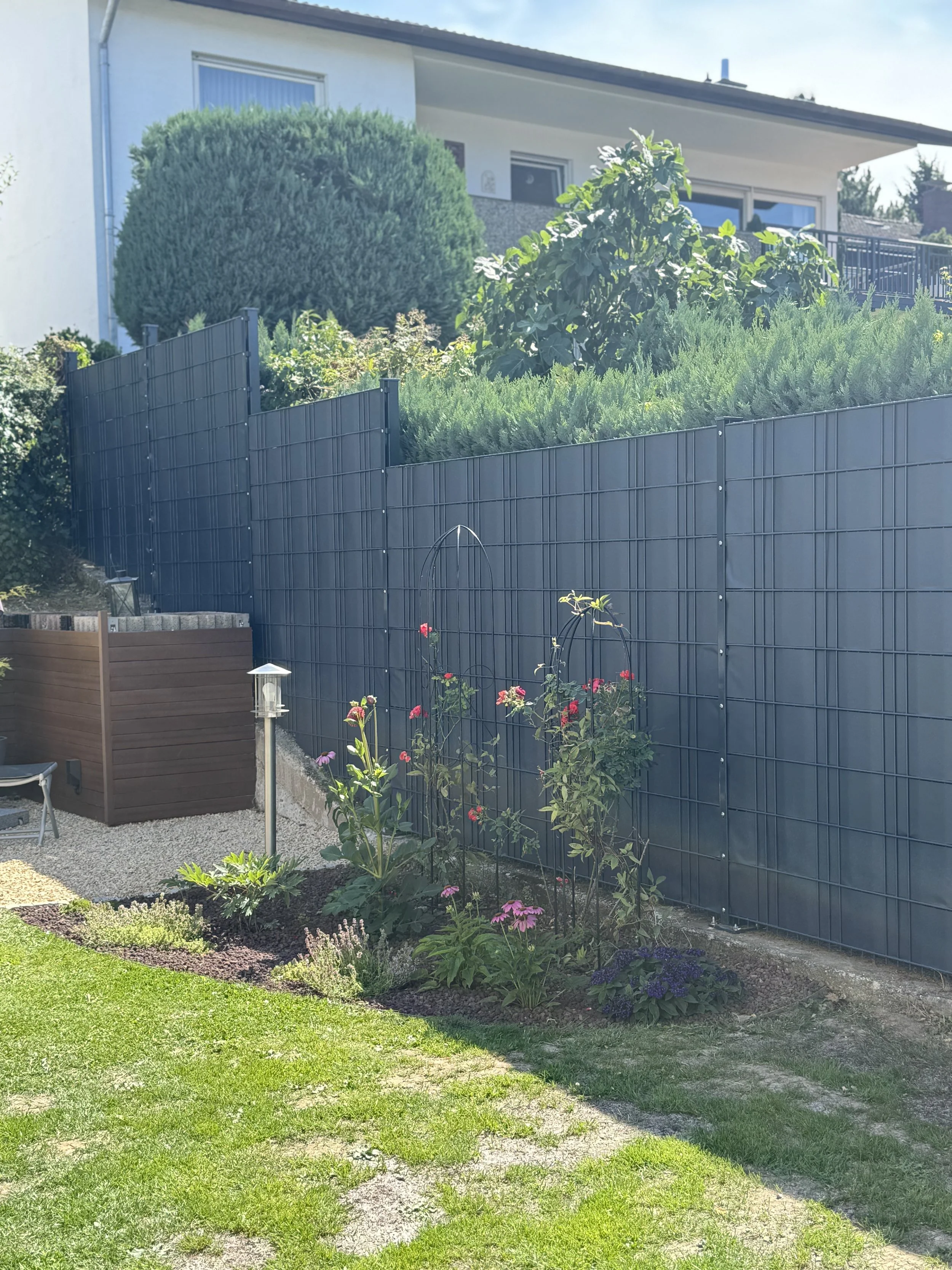 Garten mit Gräsern und Blumen vor einer schwarzen Metallfeldzaun, im Hintergrund ein Haus mit Balkon und großen Büsche.