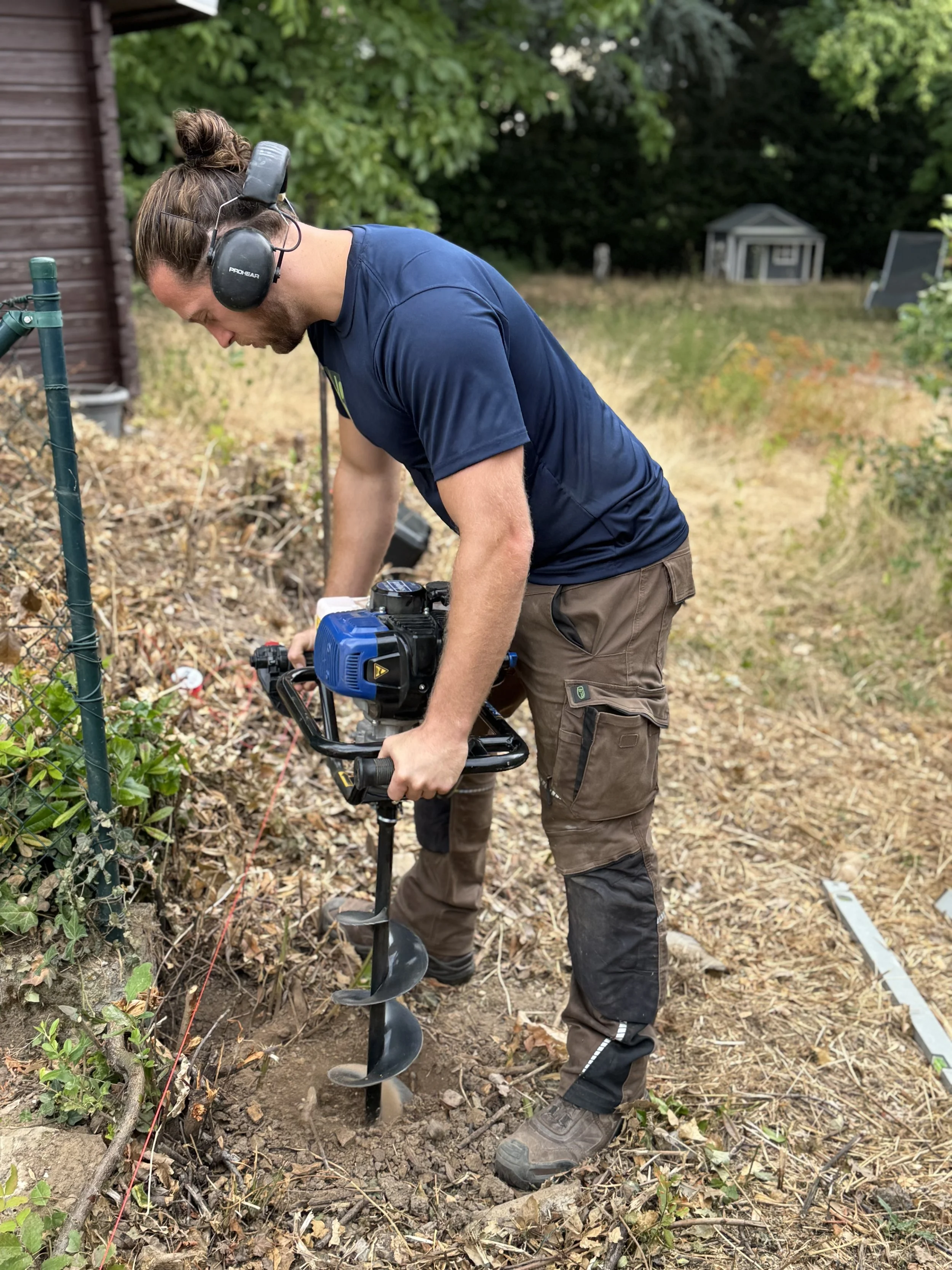 Junge männliche Person beim Bohren in den Boden mit einer Erdbohrmaschine in einem Garten mit Büschen und Bäumen.