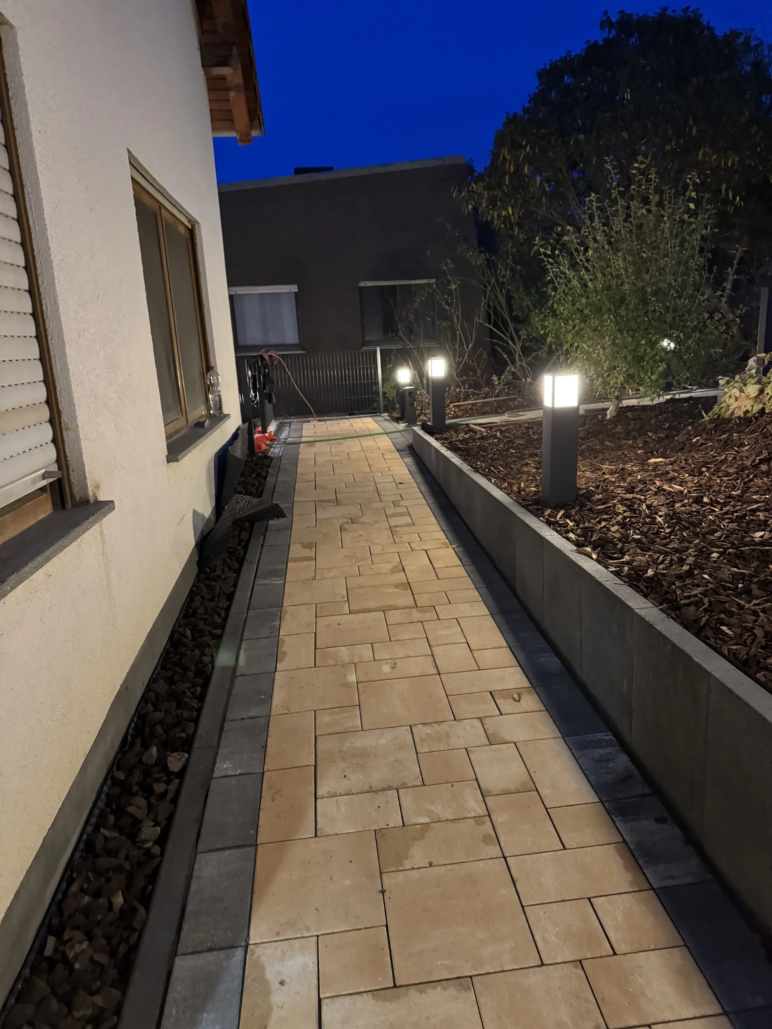 Ein beleuchteter Gartenweg mit Pflastersteinen, umgeben von dunklen Bäumen und einem Haus mit weißer Wand und Fenstern in der Abenddämmerung.