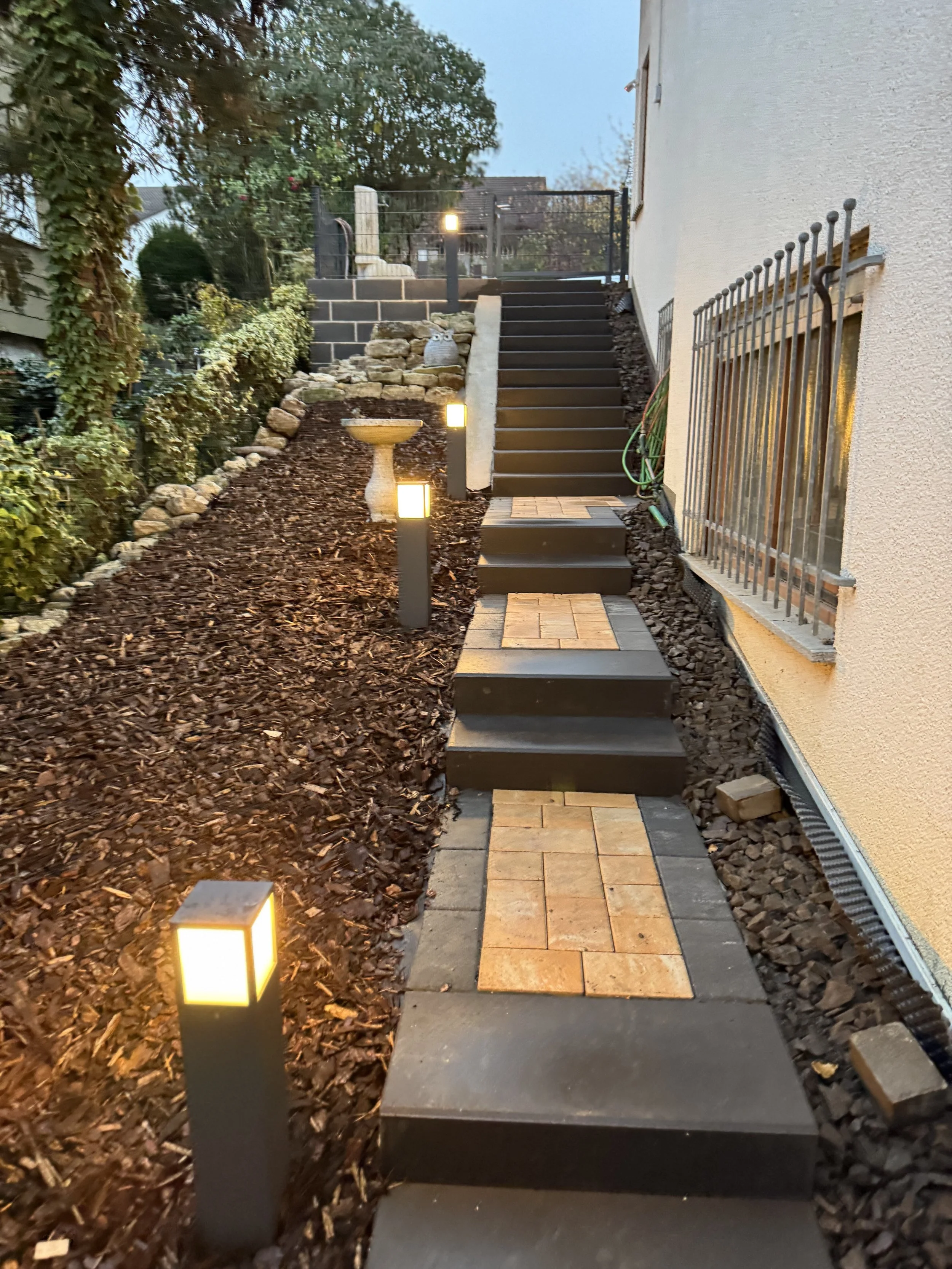 Ein gepflegter, beleuchteter Gartenweg mit Steinstufen und Laternen bei Dämmerung, umgeben von Büschen und einer Hauswand mit Fenster.