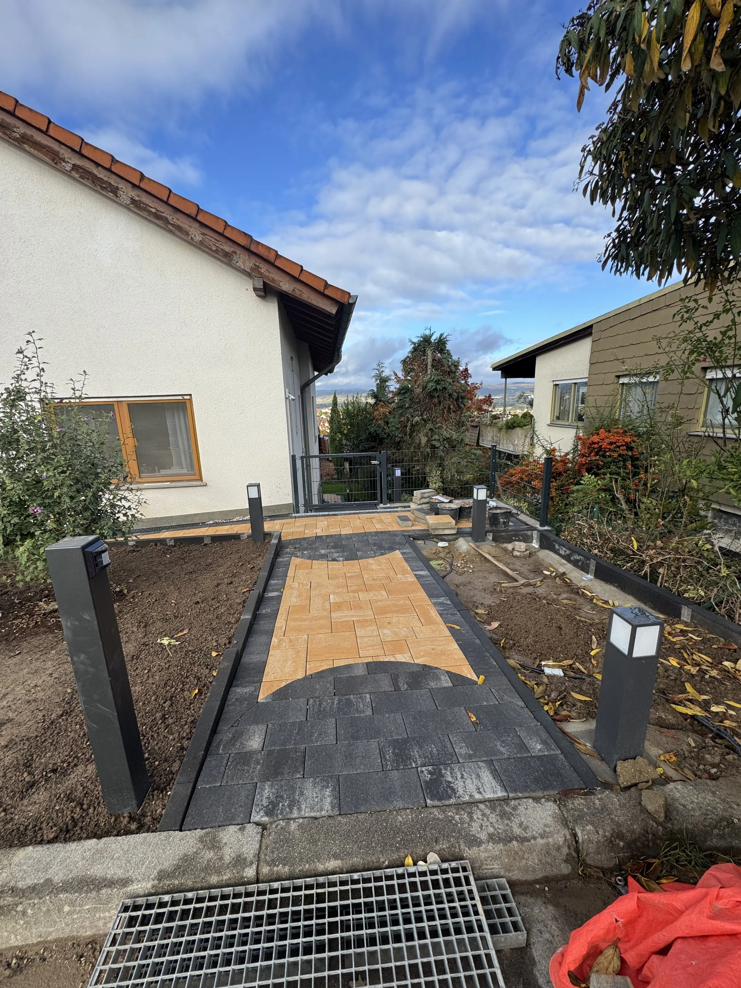 Ein frisch gepflasterter Gehweg mit schwarz-braunen Fliesen in einem Garten, umgeben von unbepflanzten Beeten, und einem Haus im Hintergrund, bei bedecktem Himmel.