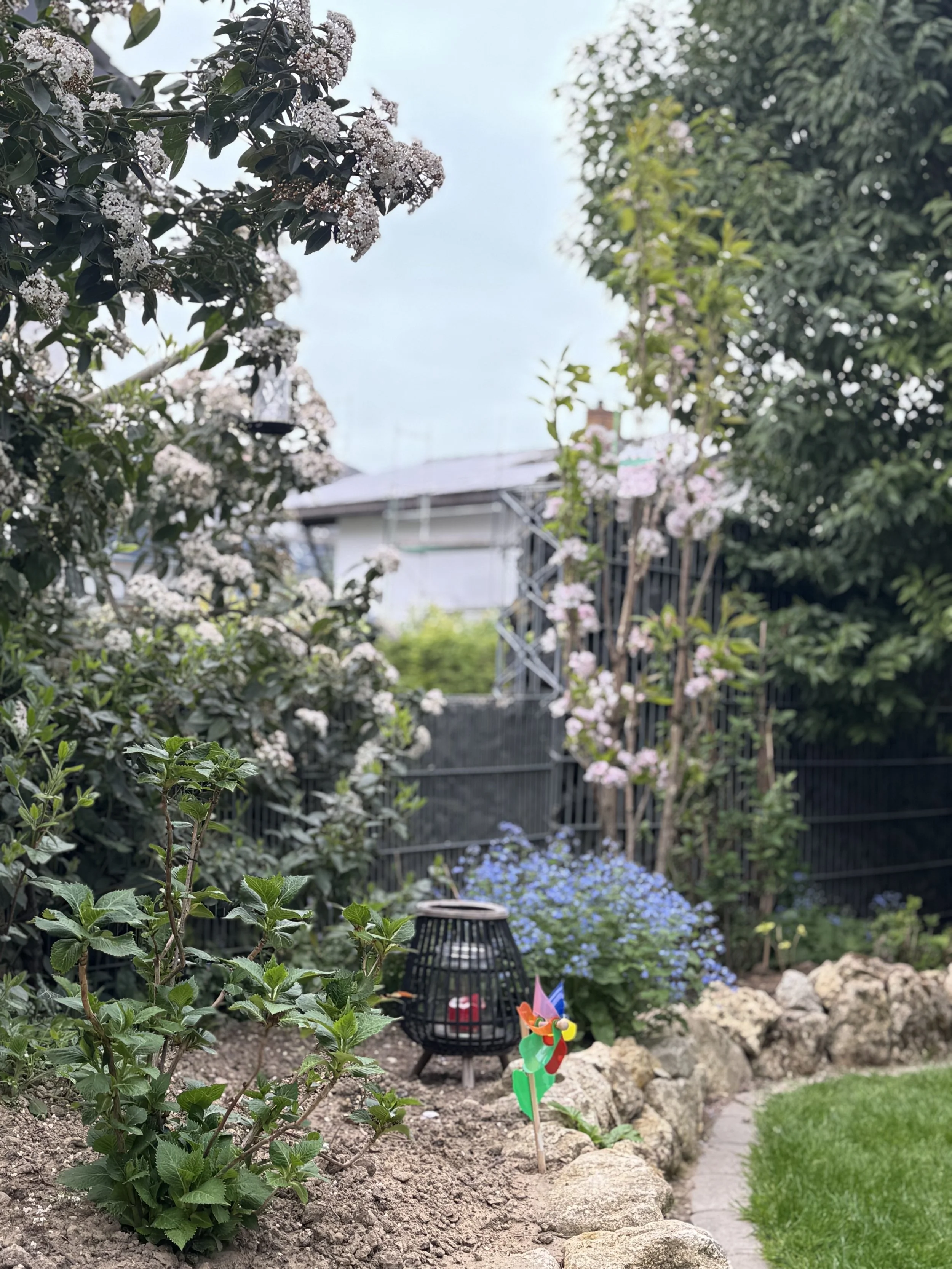 Garten mit Blumen, einem kleinen Windlicht und einer bunten Windmühle. Es gibt einen Minibach mit Steinen und eine grüne Rasenfläche.