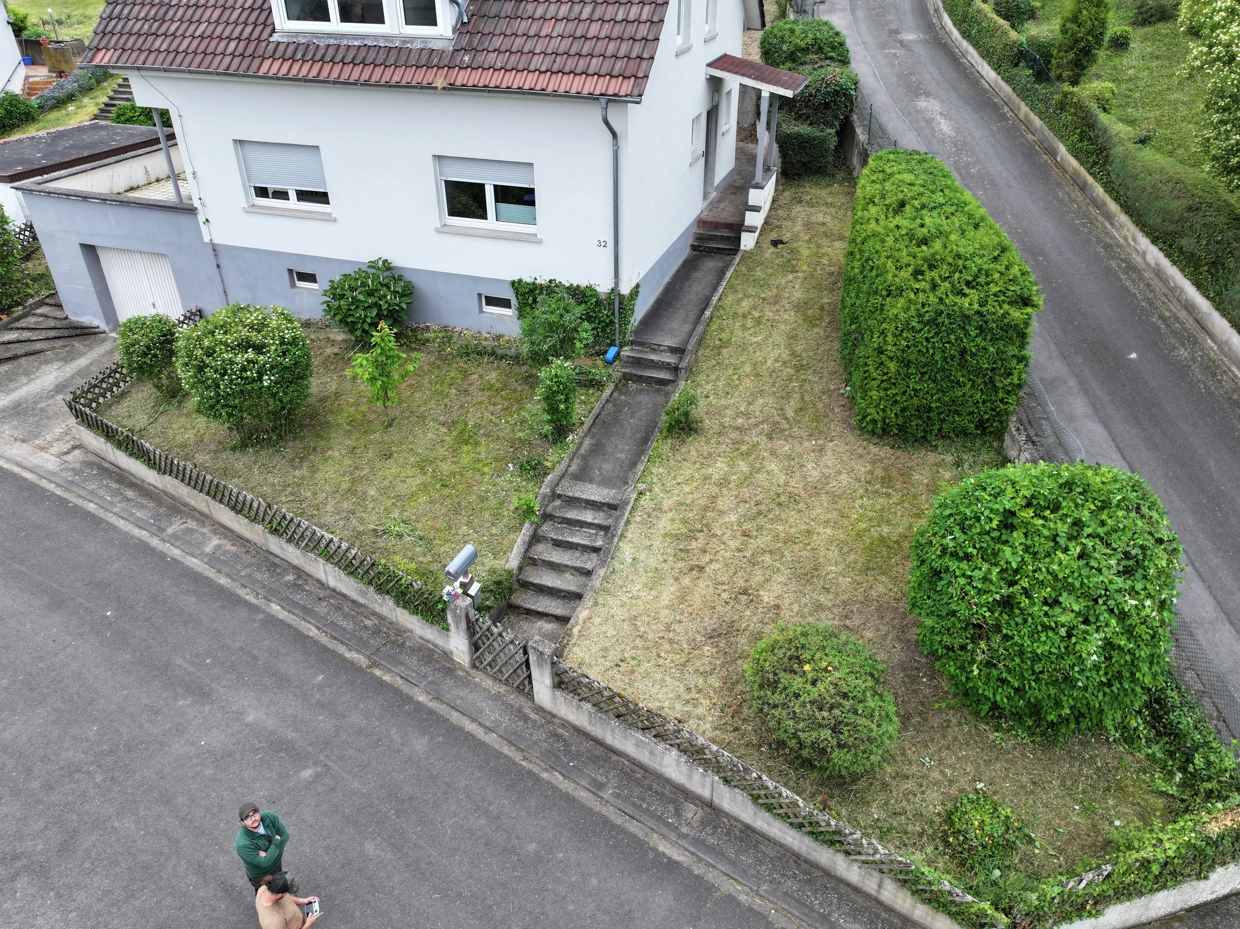 Ein weißes Wohnhaus mit roten Dach, umgeben von einem Garten mit Büschen und Bäumen, und einer Straße daneben. Im Vordergrund steht eine Person mit dunklem Shirt und Brille, die auf etwas schaut.