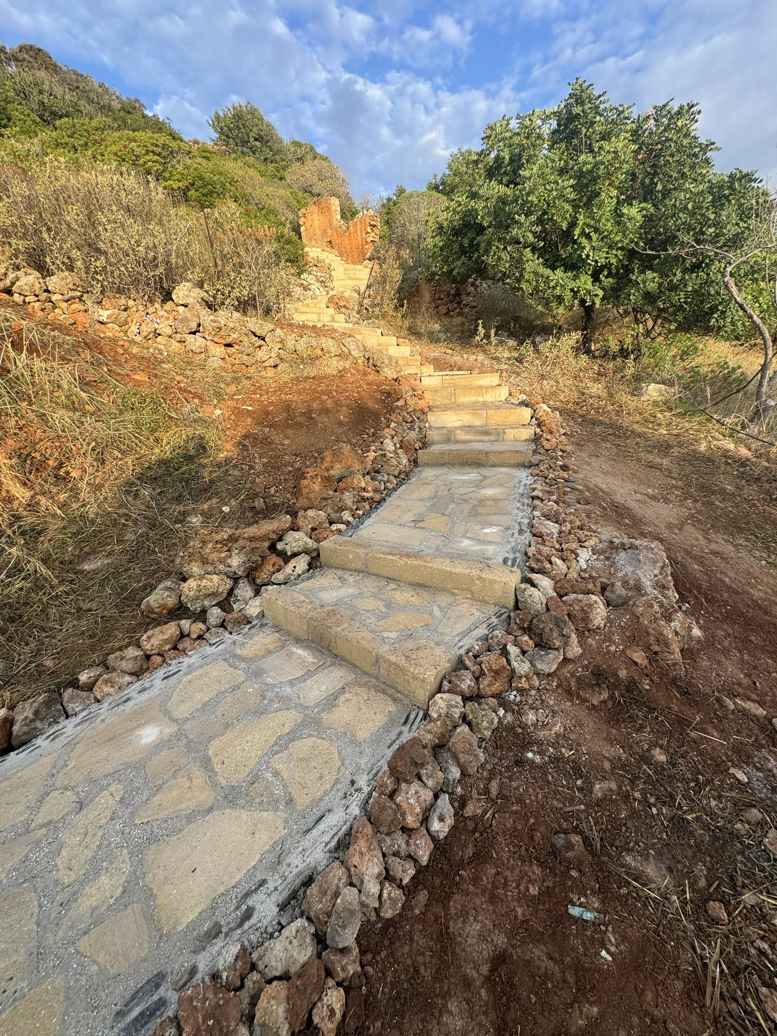 Blick auf eine neue Steintreppe, die einen schmalen Wanderweg an einem Hang hinaufführt, umgeben von trockenem Boden, Steinen, Büsche und einem Baum, mit einem blauen Himmel mit Wolken im Hintergrund.