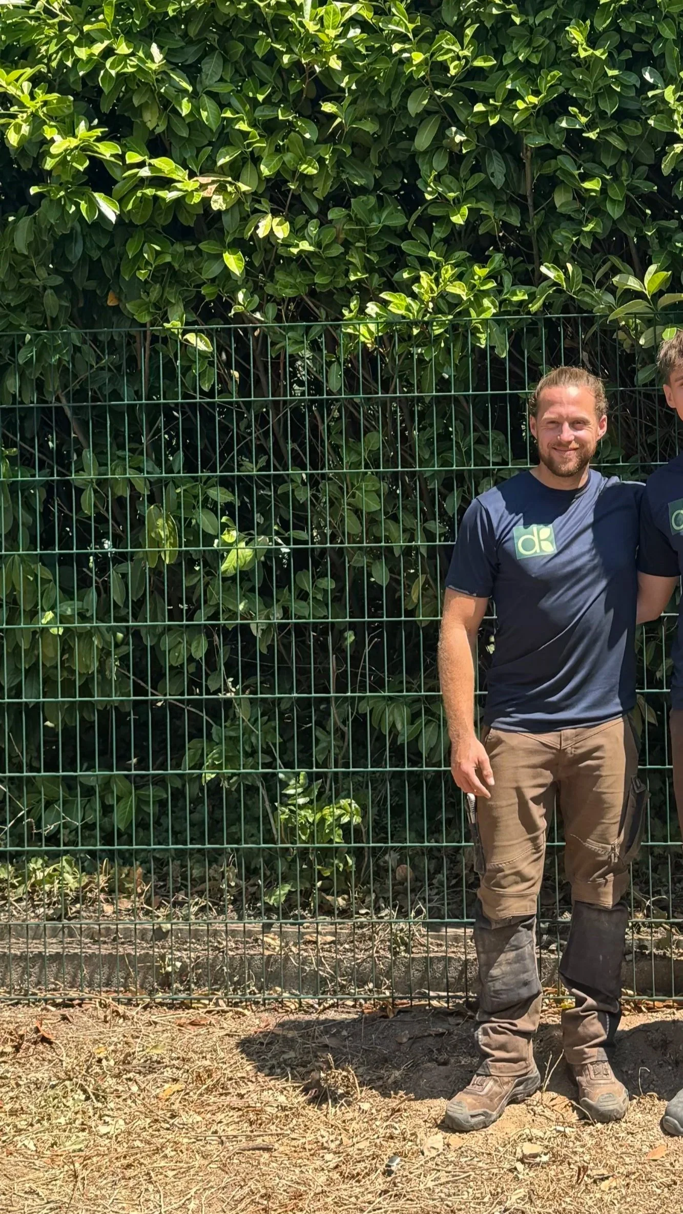 Ein Mann steht vor einem grünen Zaun, im Hintergrund dichter Laubbaum, sonniger Tag.