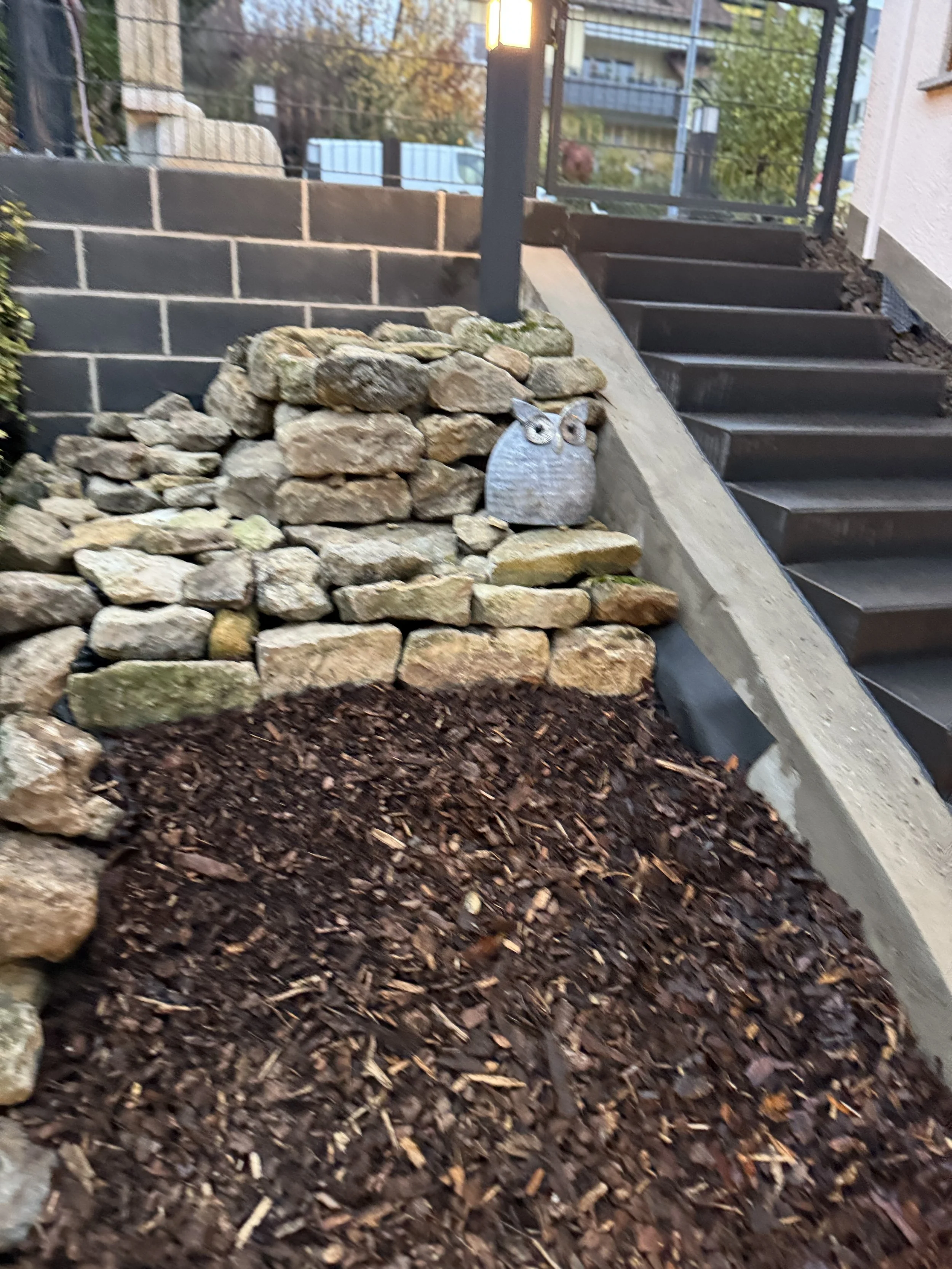 Garten mit einer Steinschale in Form eines Eulenkopfes, einem Steingarten, und einer Treppe vor einem Haus, im Hintergrund Bäume und Gebäude.