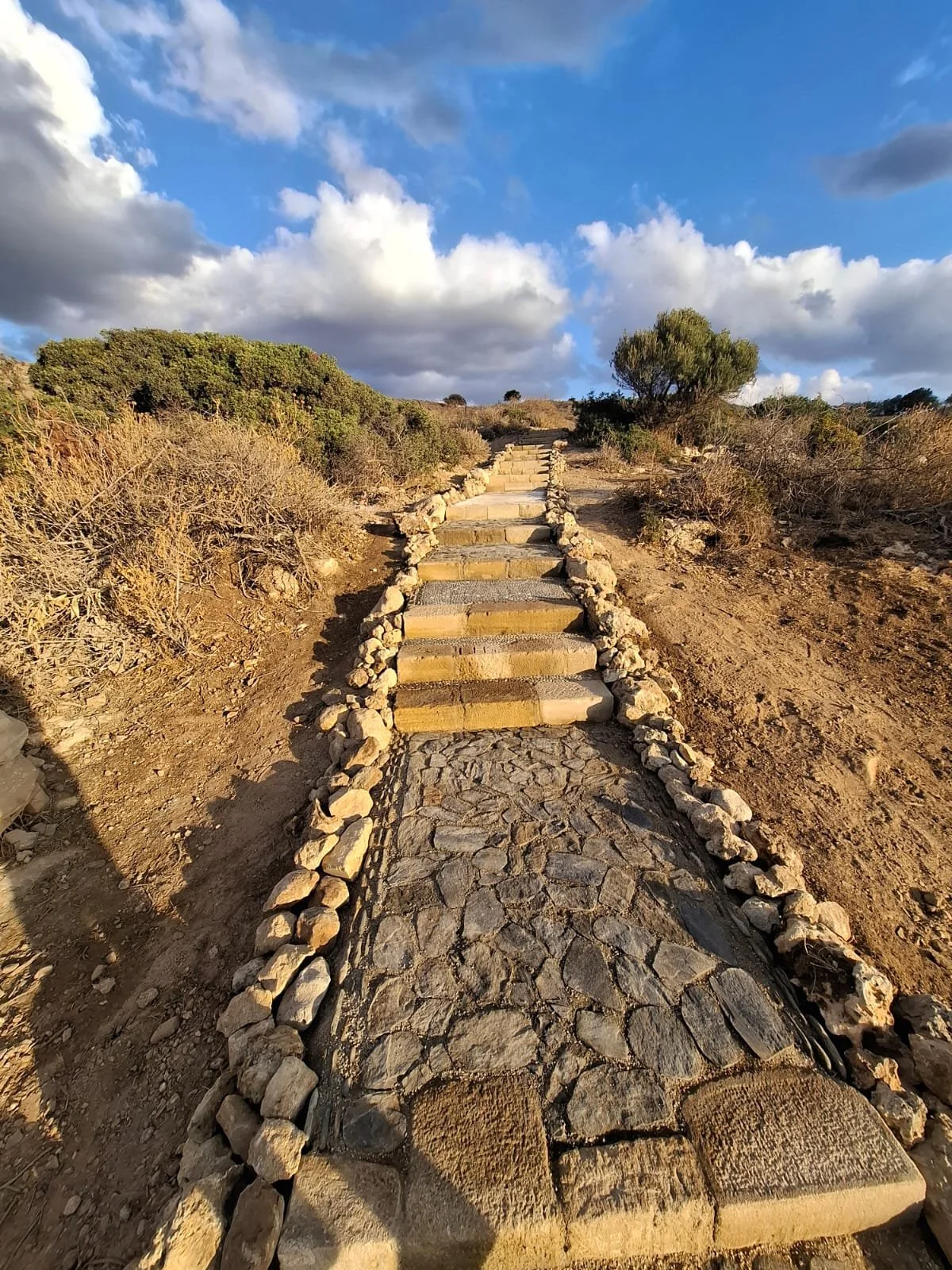 Steinpfad mit Stufen auf einem Hügel, umgeben von trockener Vegetation unter einem blauen Himmel mit Wolken.
