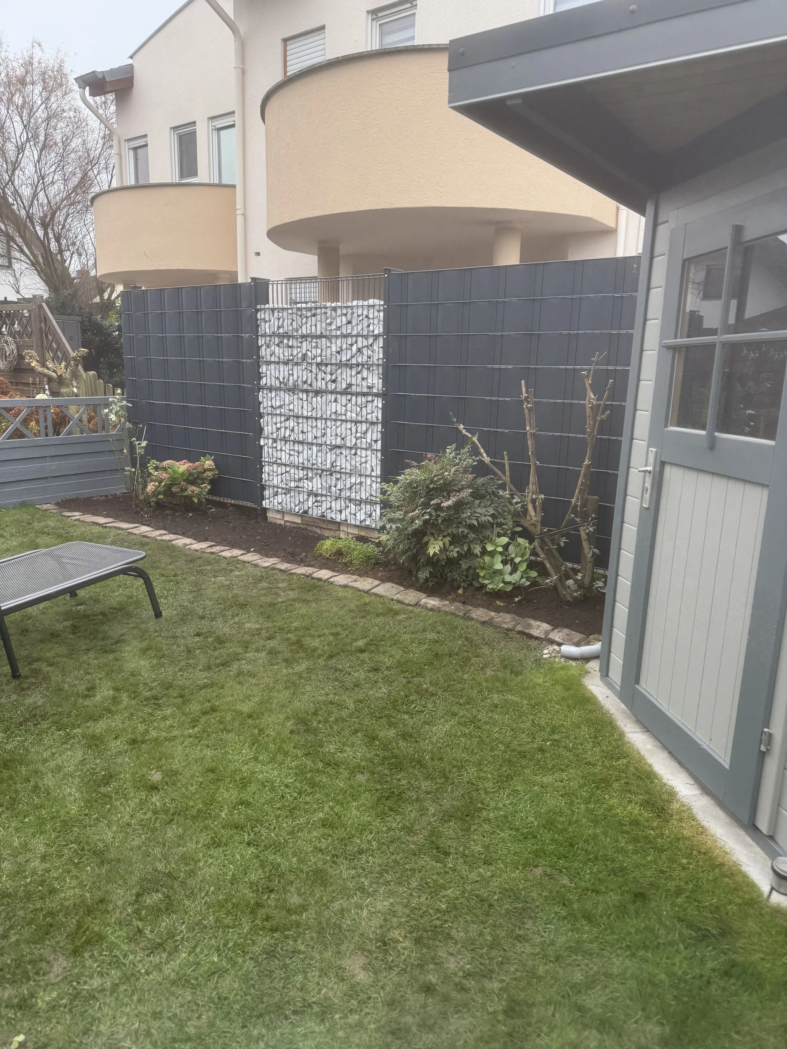 Garten mit grünem Rasen, Blumenbeeten und einem schwarzen Gartensessel, eingezäunt mit einer schwarzen Metallzaun und einer dekorativen Wand aus weißen Steinen, neben einem Gartenhäuschen.