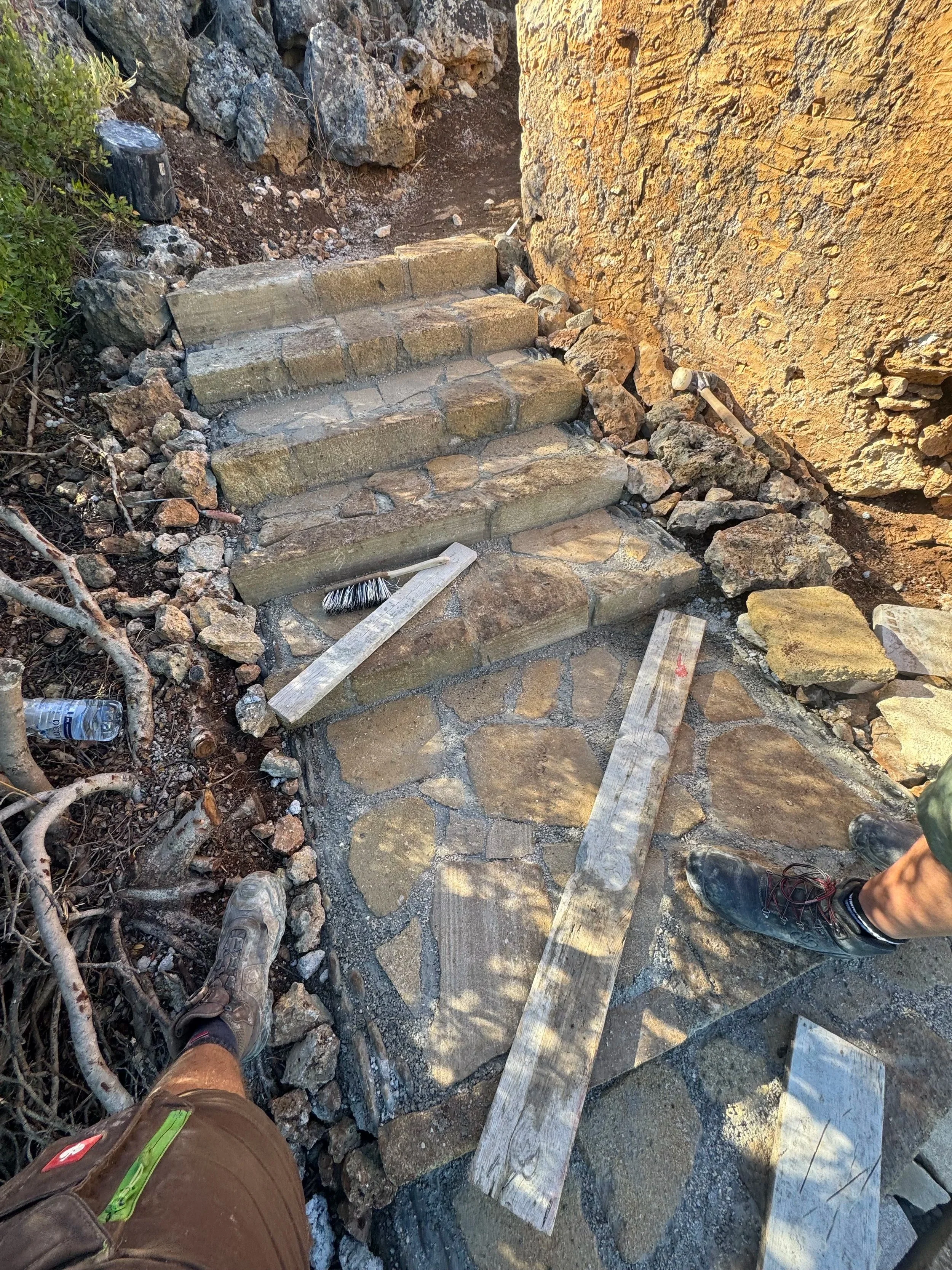 Bauarbeiten an einer Stein- und Ziegelstufe im Freien, mit Bauwerkzeugen und zwei Personen in Wanderschuhen.