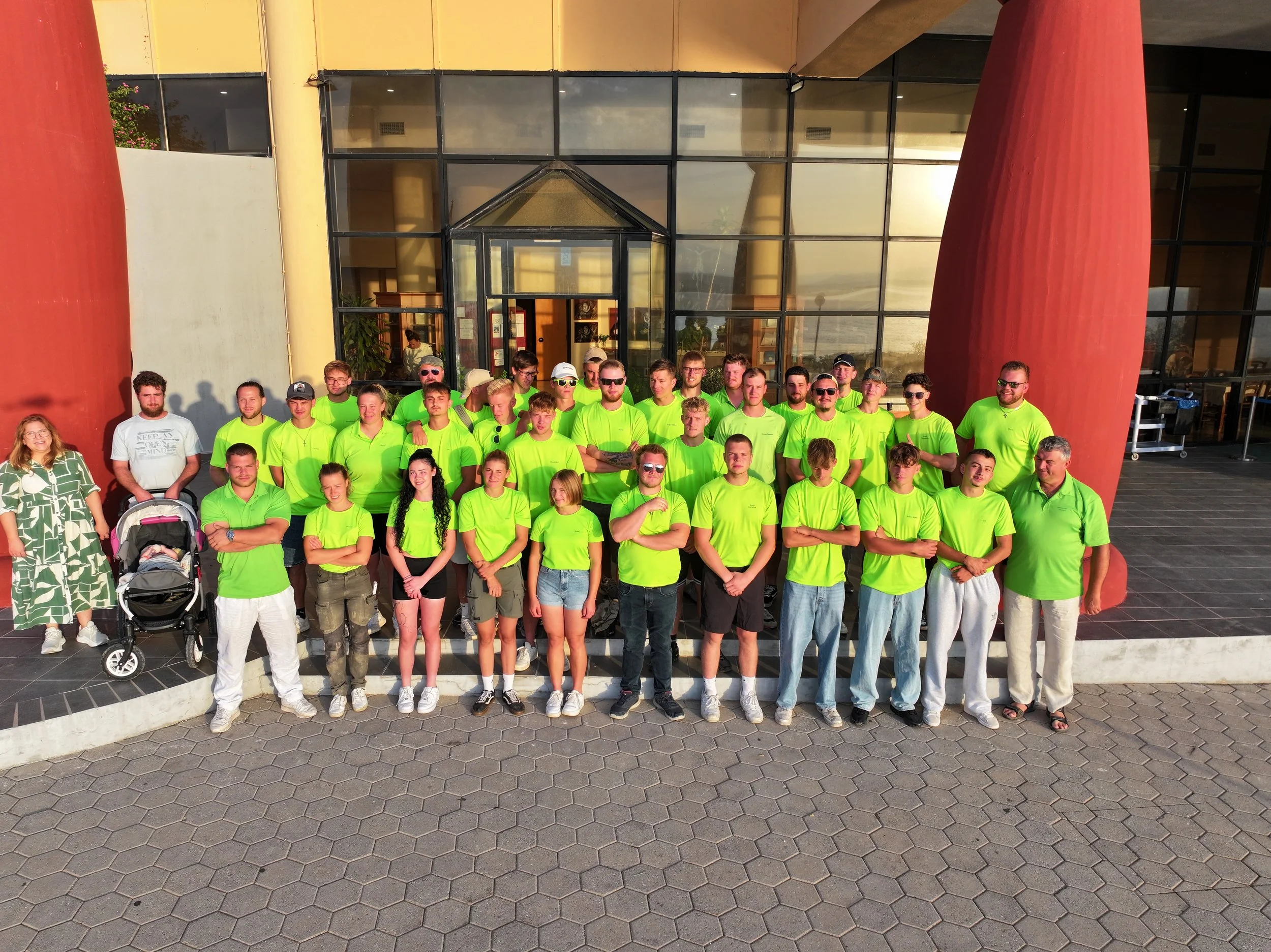 Gruppe von Menschen vor einem modernen Gebäude, viele tragen neon grüne T-Shirts, sonniges Wetter.