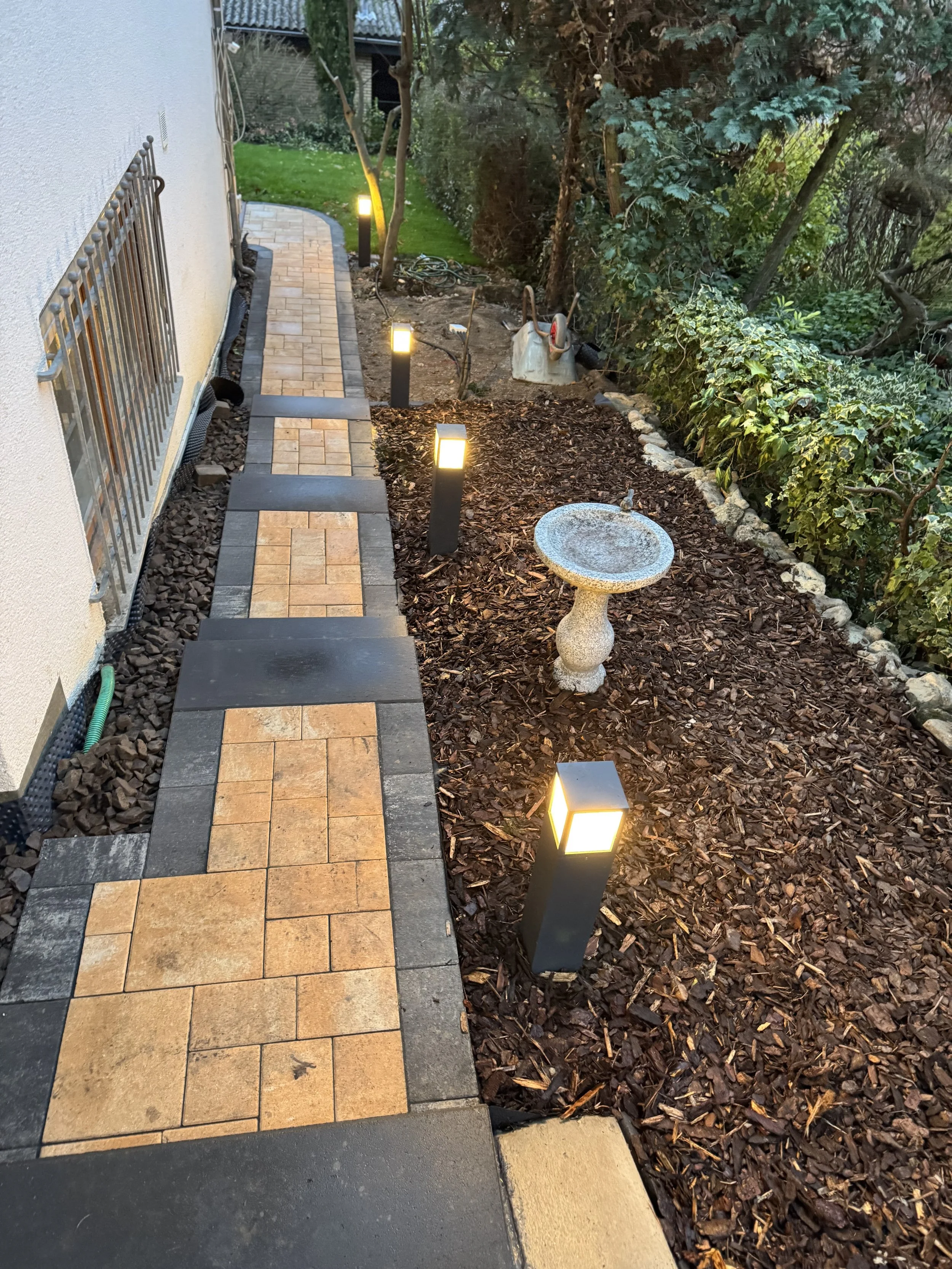 Ein Gartenweg mit Bodenplatten, seitlich vom Haus leuchtende Wegleuchten, eine Vogeltränke und Bäume im Hintergrund.