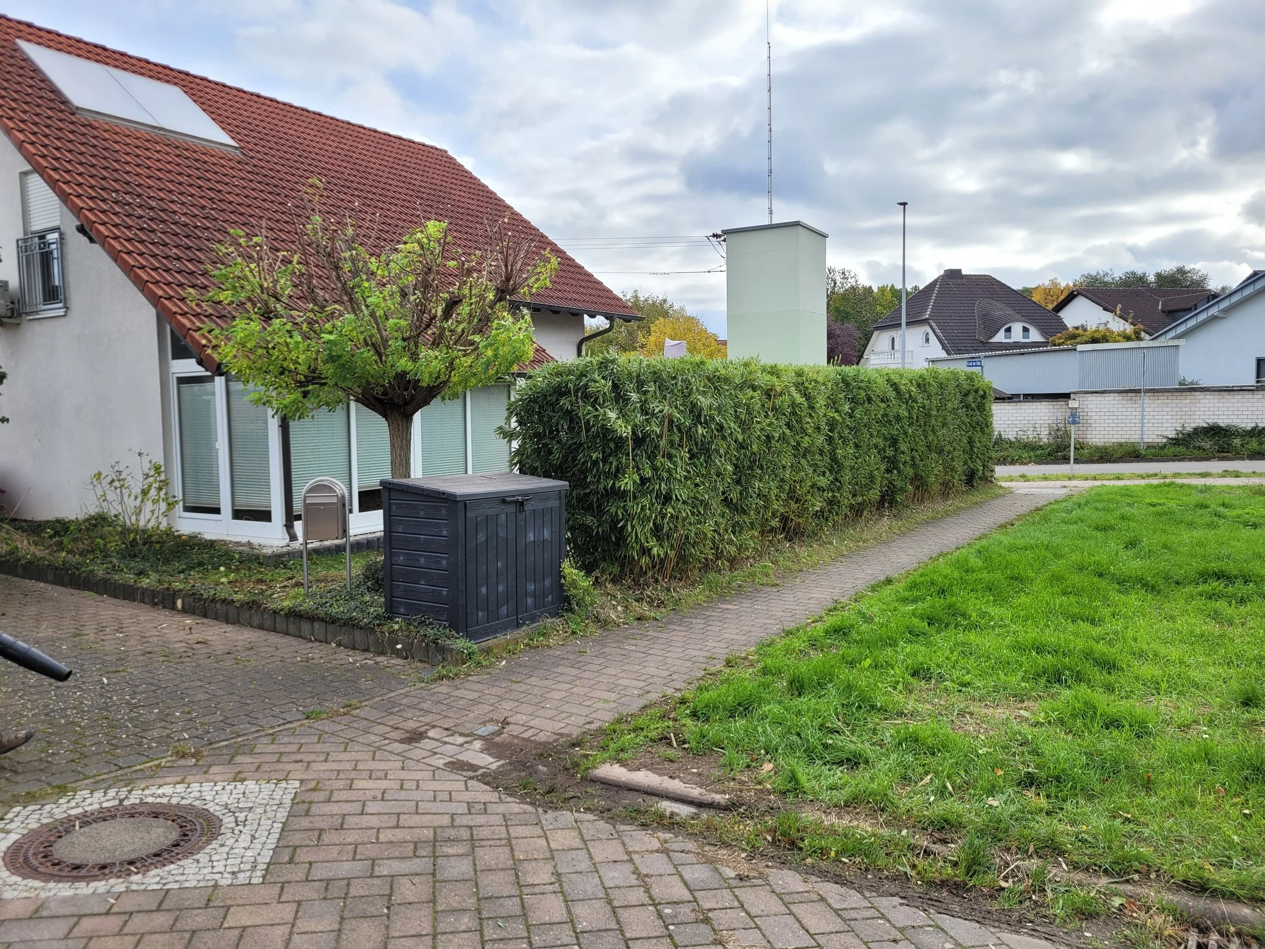 Ein Garten mit einer roten Ziegeldach-Haus, einer Baum, einer Hecke, einem schwarzen Gartenhäuschen, einem Weg, einer Wiese und umgebenden Häusern bei bewölktem Himmel.