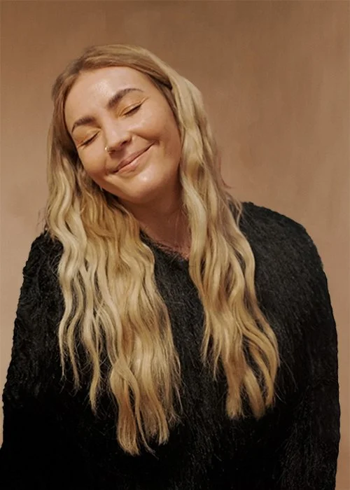 Eine lachende Frau mit langem, blondem, gewelltem Haar und dunklem Pullover vor einem beigen Hintergrund. Katharina Dietrich ist Salon Managerin, Marketingbeauftragte, Kommunikationsdesignerin im HairClub Bad Oeynhausen.