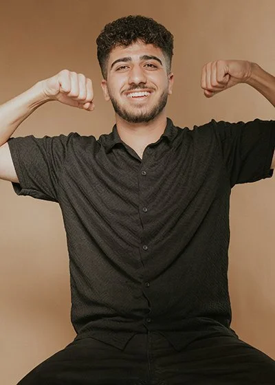 Junger Mann mit dunklem, lockigem Haar und Bart, der lächelnd in die Kamera schaut und seine gestreckten Arme zeigt, als würde er Muskeln anspannen, vor einem beigen Hintergrund. Dear Saido Shammo ist Auszubildender/Junior Stylist im HairClub.