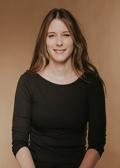 Eine junge Frau mit langem braunem, glänzendem und gesunden Haar, trägt ein schwarzes Oberteil und lächelt vor einem beigen Hintergrund. Janina Waskow ist Mitarbeiterin im HairClub Bad Oeynhausen ist Top Stylistin.