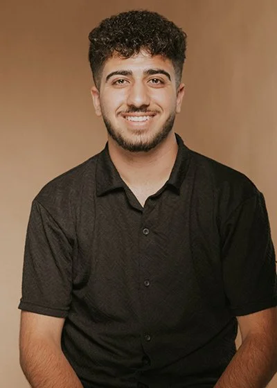 Junger Mann mit dunklem Hemd vor neutralem Hintergrund, lächelnd. Dear Saido Shammo ist Junior Stylist, Auszubildender Friseur im Friseursalon HairClub in Bad Oeynhausen.