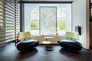 Sitzbereich mit zwei schwarzen Designersesseln von Newyesterday (Händler für Vintage Möbel), einem kleinen runden Tisch, einer großen Fensterwand mit Blick nach draußen, und einer weißen Jalousie im HairClub Friseur Salon in Bad Oeynhausen.