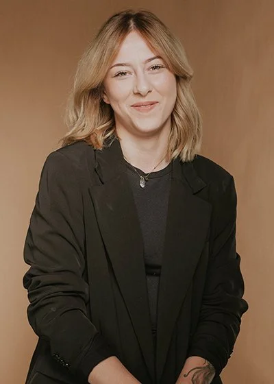 Junge Frau mit Schulterlänge blondem Haar, schwarzem Blazer und Kette, vor braunem Hintergrund. Emily Schindler ist Top Stylistin, professionelle Friseurin im HairClub Bad Oeynhausen und auf professionelle Haarfärbetechniken spezialisiert.