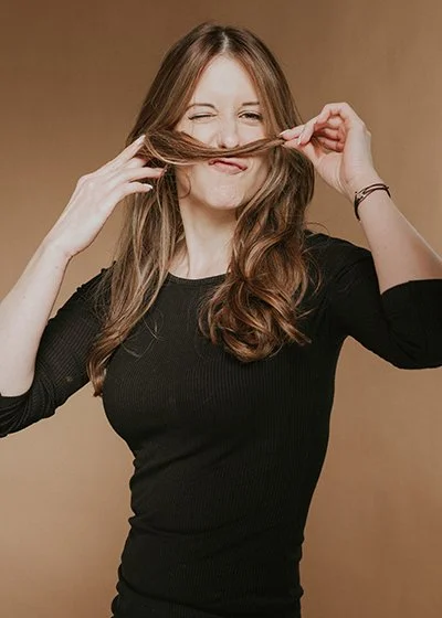 Junge Frau mit sehr gesunden braunen Haaren, die sich die Haare vor dem Gesicht hält und eine verspielte Miene macht. Janina Waskow ist Mitarbeiterin im HairClub Bad Oeynhausen ist Top Stylistin.